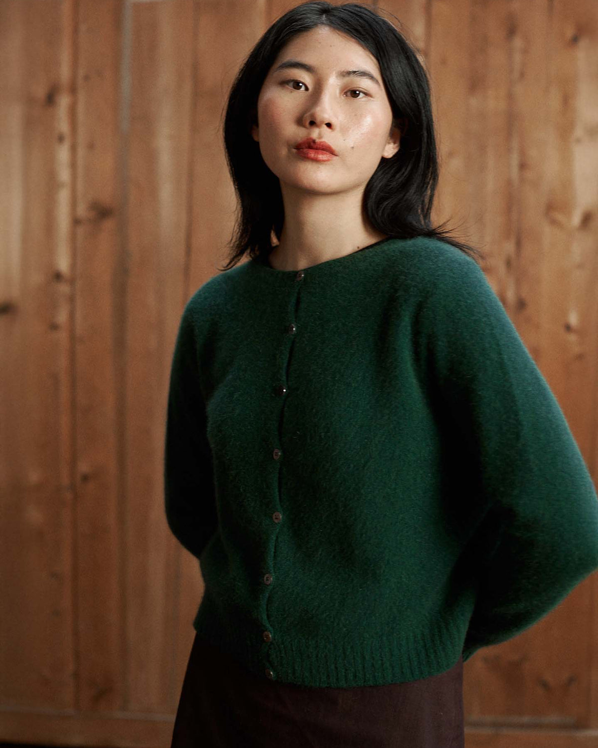 A woman with medium-length dark hair wears the Dusk Cardigan in Verdant Green, a relaxed fit buttoned sweater, as she stands before a wooden wall and looks at the camera.