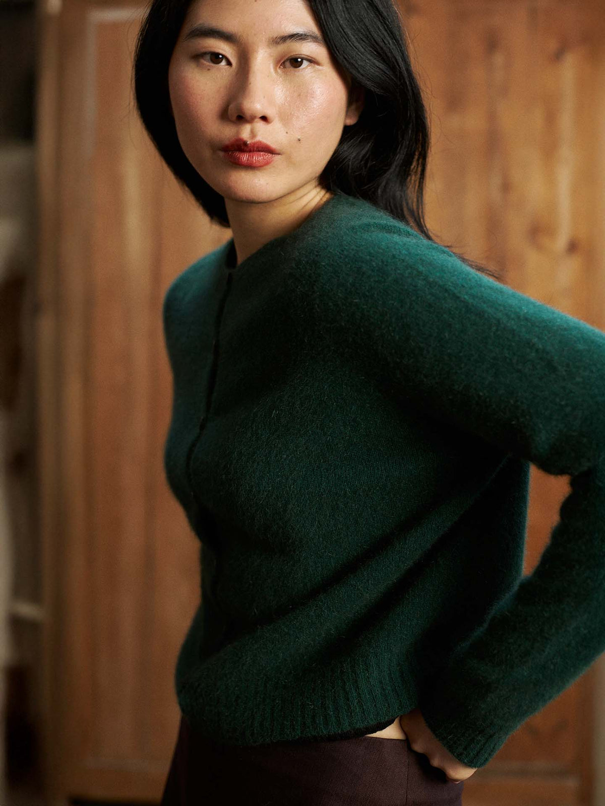 A woman with long black hair wears the Dusk Cardigan in Verdant Green, a relaxed-fit fuzzy sweater, standing indoors against a wooden background and looking toward the camera.