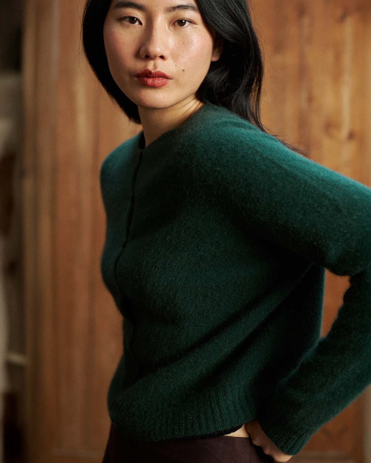 A woman with long black hair wears the Dusk Cardigan in Verdant Green, a relaxed-fit fuzzy sweater, standing indoors against a wooden background and looking toward the camera.