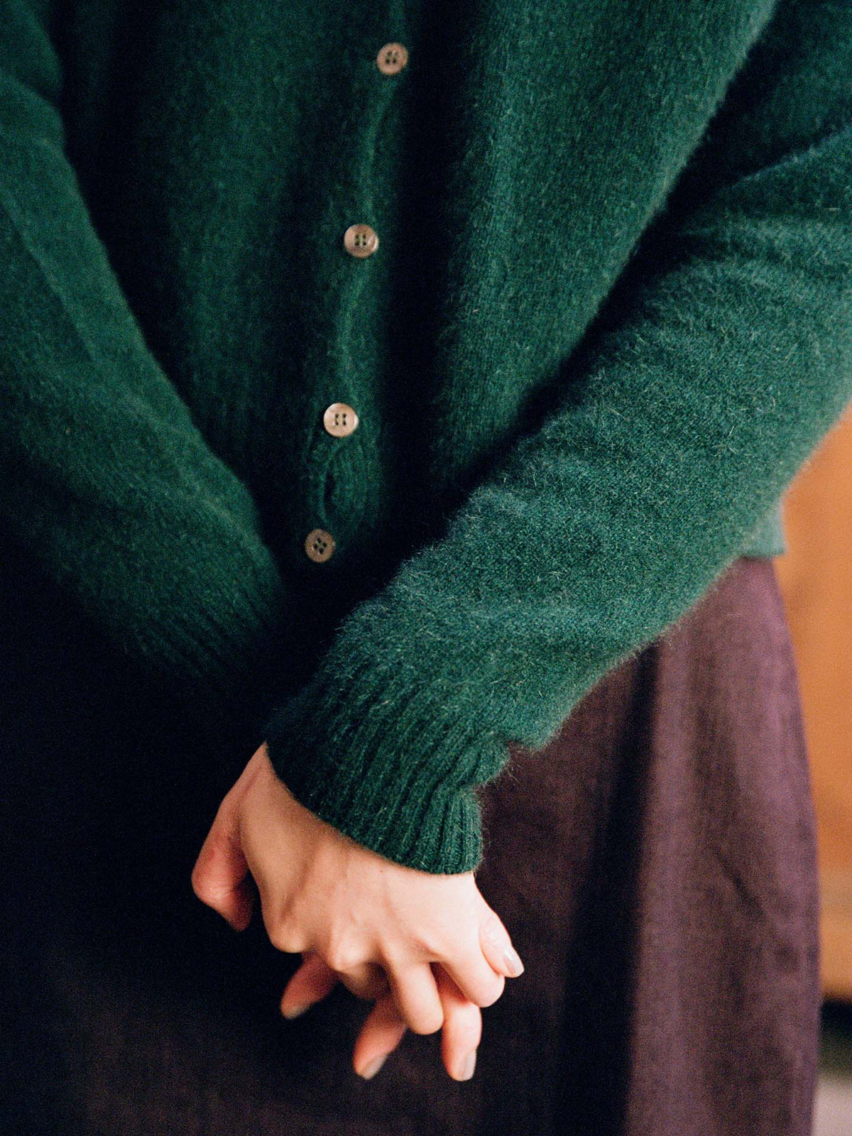 A person wearing the Dusk Cardigan in Verdant Green with a relaxed fit and a dark skirt stands with hands clasped at their waist.