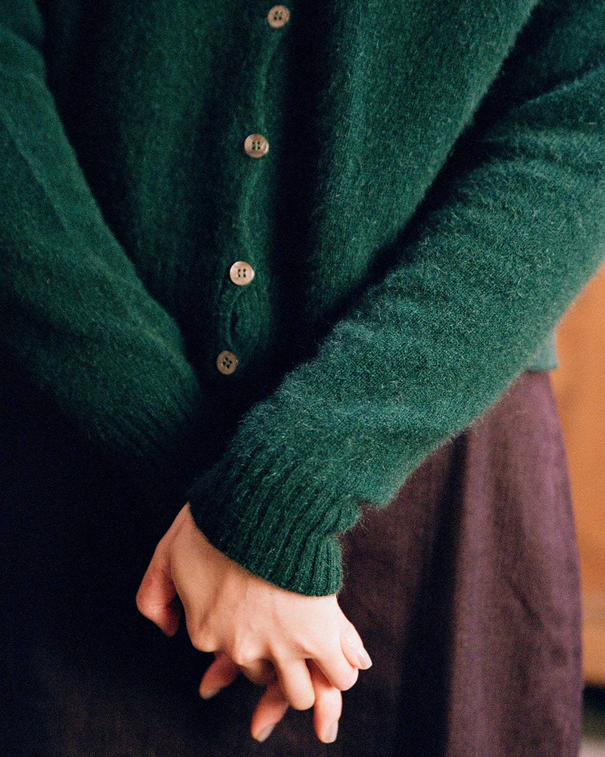 A person wearing the Dusk Cardigan in Verdant Green with a relaxed fit and a dark skirt stands with hands clasped at their waist.