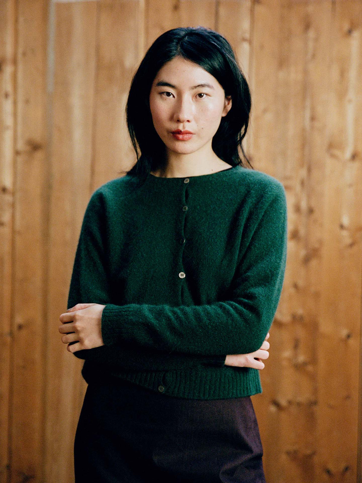 A person with long dark hair wears the Dusk Cardigan in Verdant Green and a dark skirt, standing before a wooden wall and looking at the camera with arms loosely crossed.