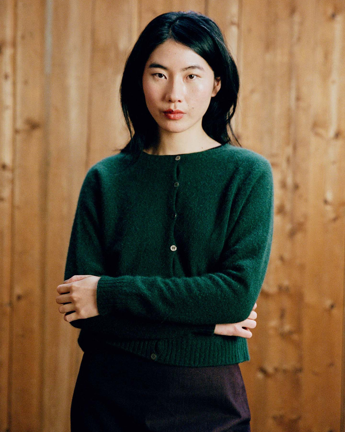 A person with long dark hair wears the Dusk Cardigan in Verdant Green and a dark skirt, standing before a wooden wall and looking at the camera with arms loosely crossed.