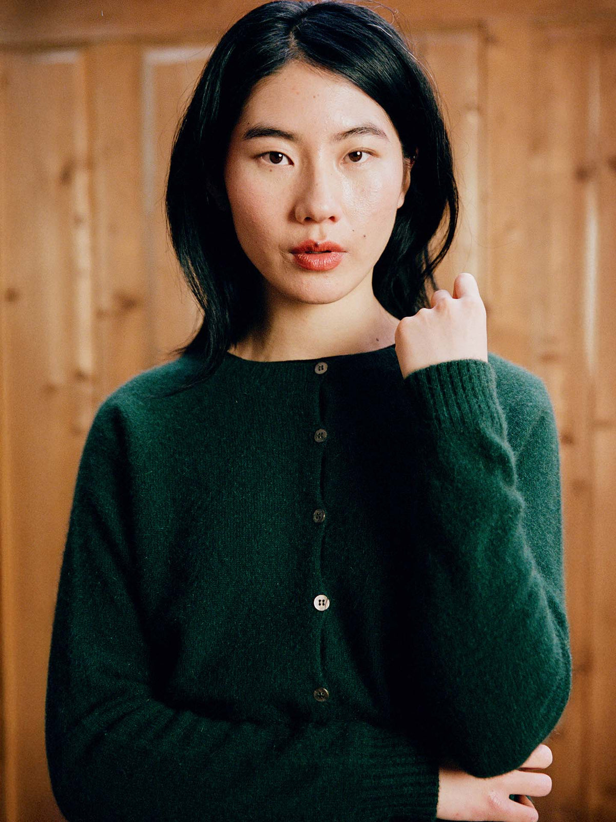 A person with long black hair wears the Dusk Cardigan in Verdant Green, a relaxed fit button-up sweater, and stands before a wooden background, looking directly at the camera.