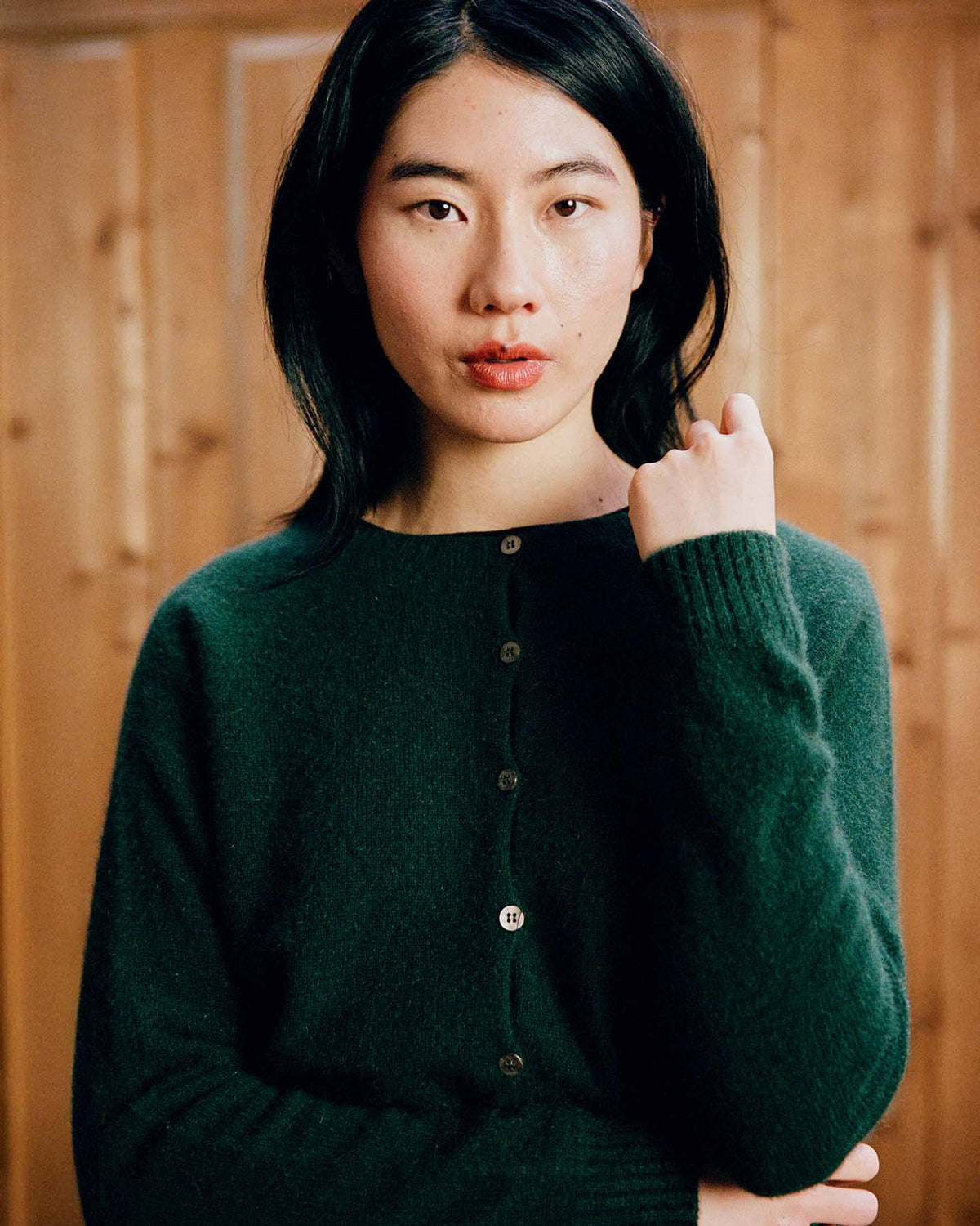 A person with long black hair wears the Dusk Cardigan in Verdant Green, a relaxed fit button-up sweater, and stands before a wooden background, looking directly at the camera.
