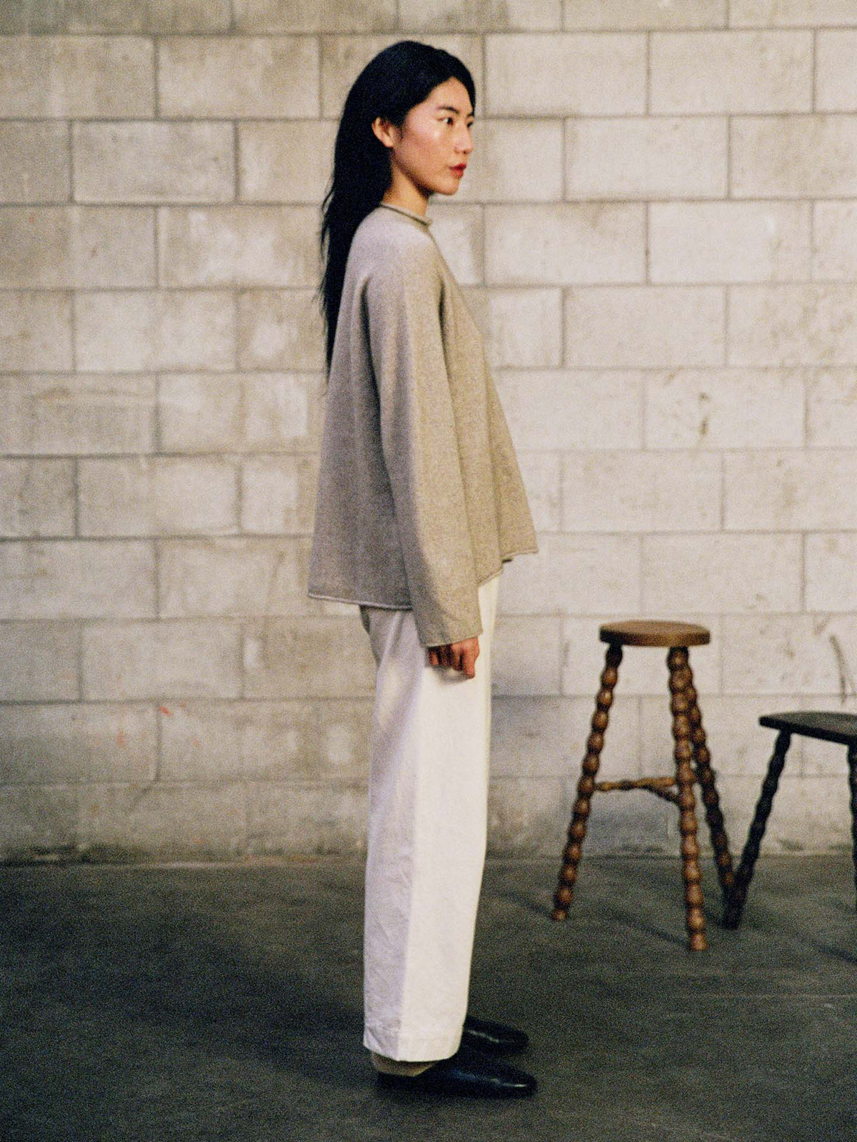 A woman in profile stands before a cinder block wall, dressed in the oversized Eclipse Knit – Natural sweater with white pants and black shoes. Two wooden stools are visible behind her.