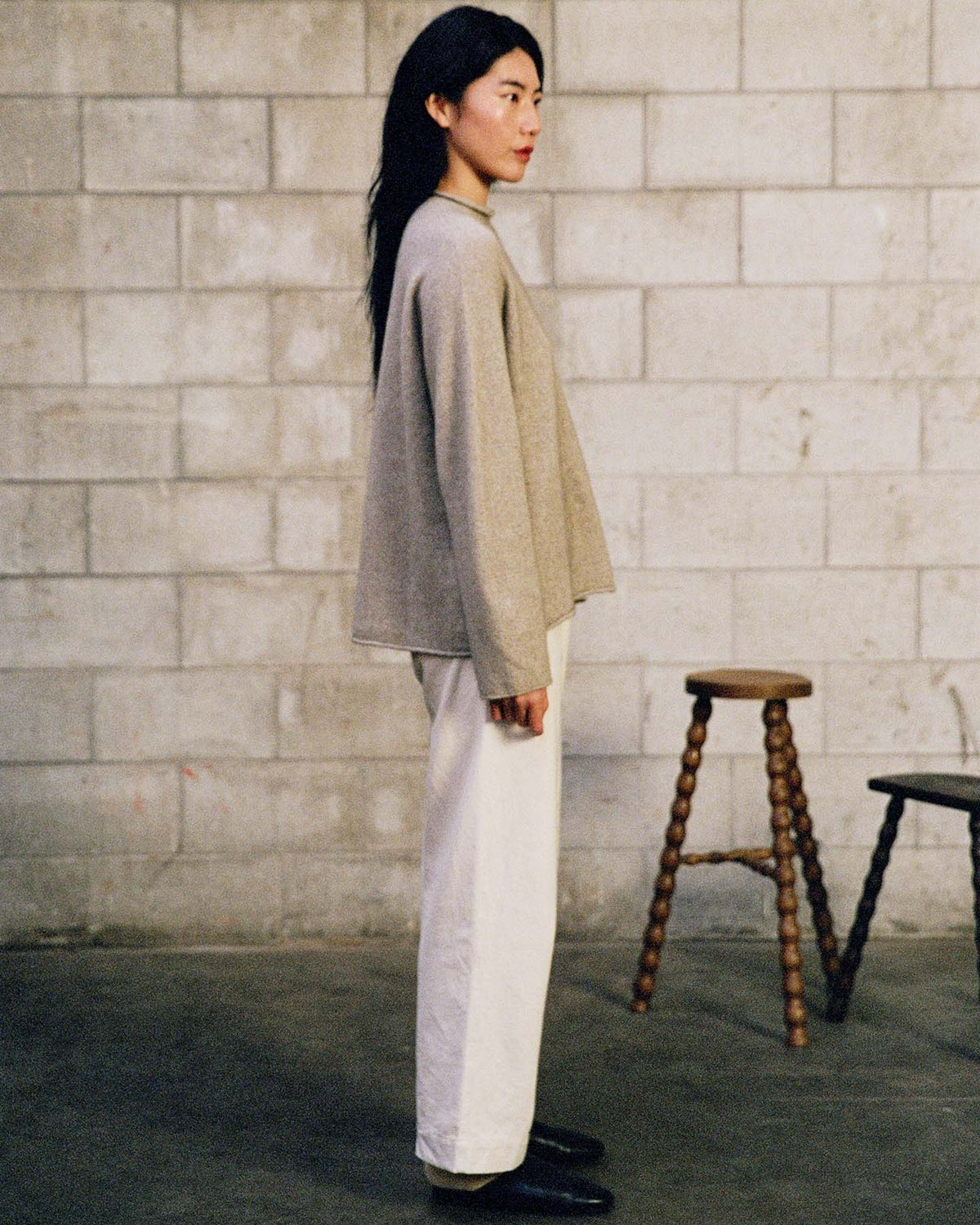 A woman in profile stands before a cinder block wall, dressed in the oversized Eclipse Knit – Natural sweater with white pants and black shoes. Two wooden stools are visible behind her.