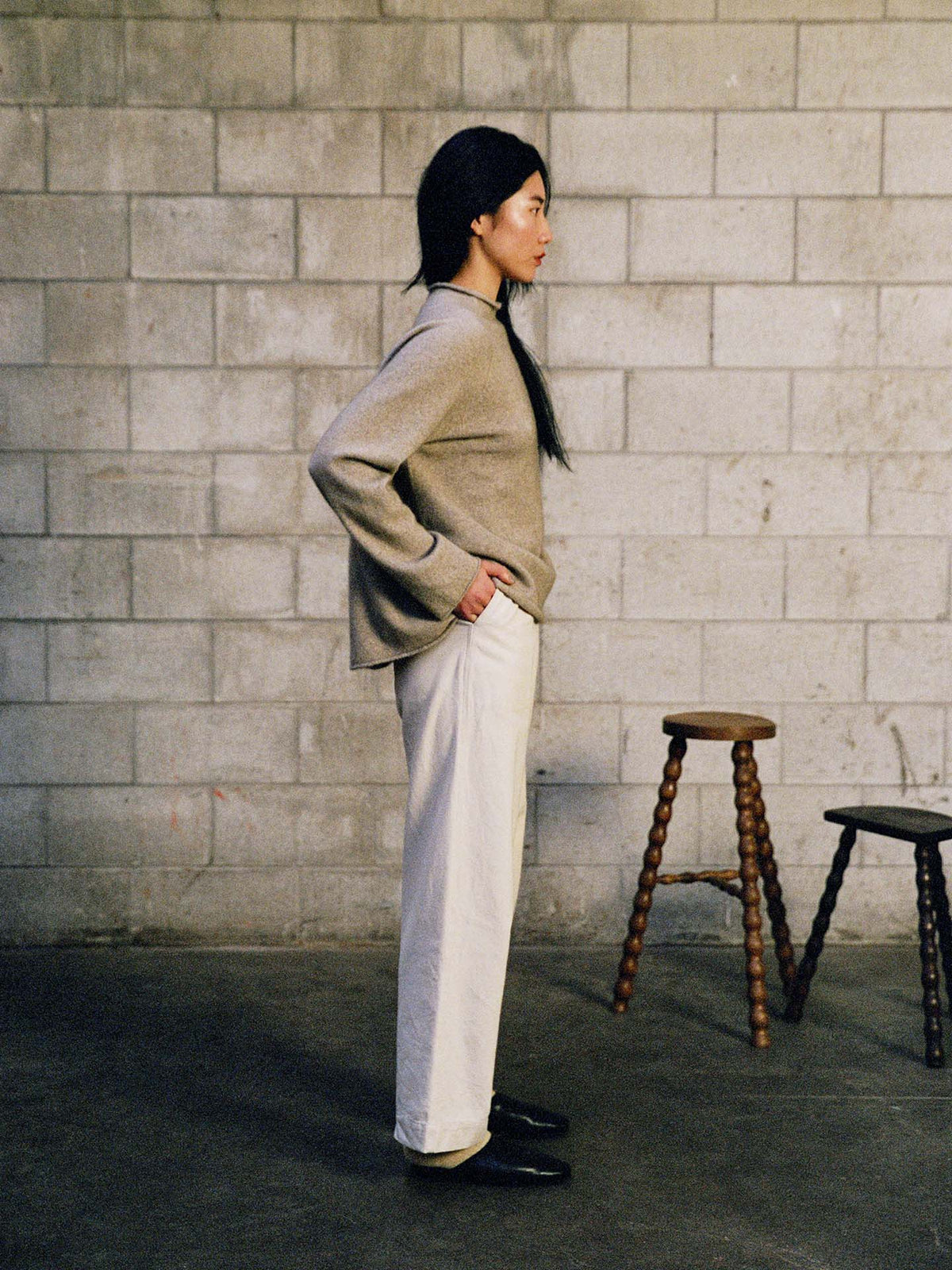 A person stands in profile against a concrete block wall, wearing the Eclipse Knit – Natural sweater with white pants and black shoes, while two wooden stools are nearby.