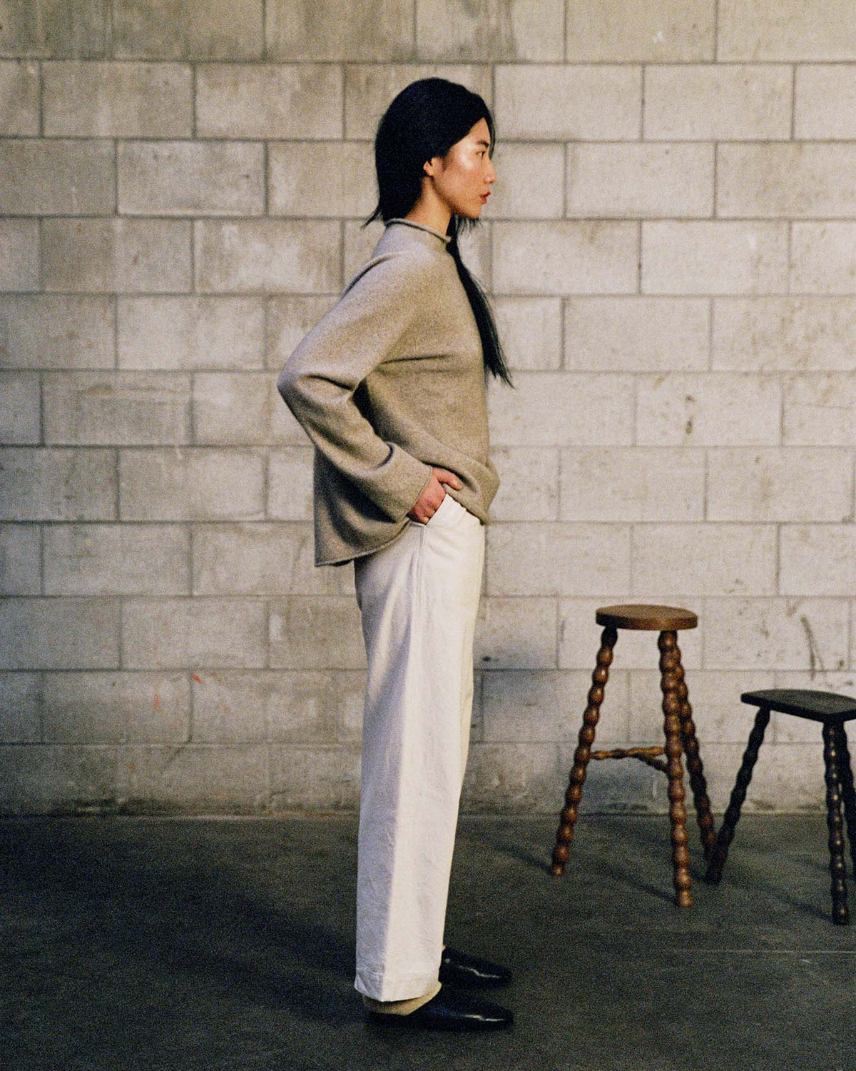 A person stands in profile against a concrete block wall, wearing the Eclipse Knit – Natural sweater with white pants and black shoes, while two wooden stools are nearby.