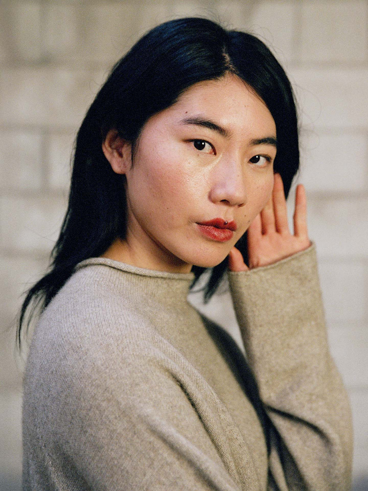 A woman with long dark hair and a neutral expression poses indoors, wearing the Eclipse Knit – Natural, an oversized light gray sweater, with one hand near her ear.