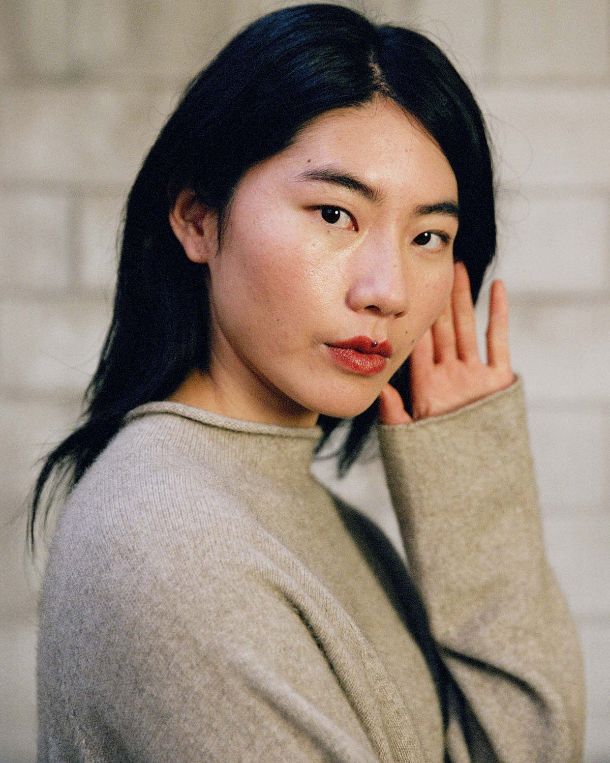 A woman with long dark hair and a neutral expression poses indoors, wearing the Eclipse Knit – Natural, an oversized light gray sweater, with one hand near her ear.