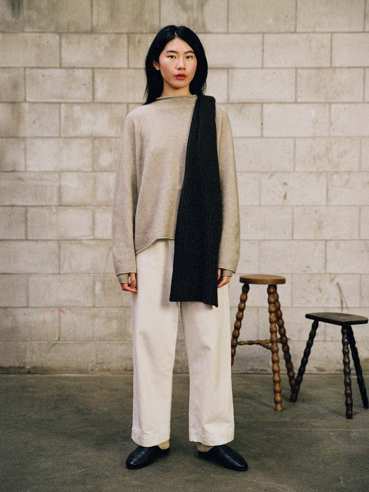 A person stands indoors against a concrete block wall, wearing the Eclipse Knit – Natural sweater, white pants, black shoes, and a black scarf. Two wooden stools are positioned behind them.