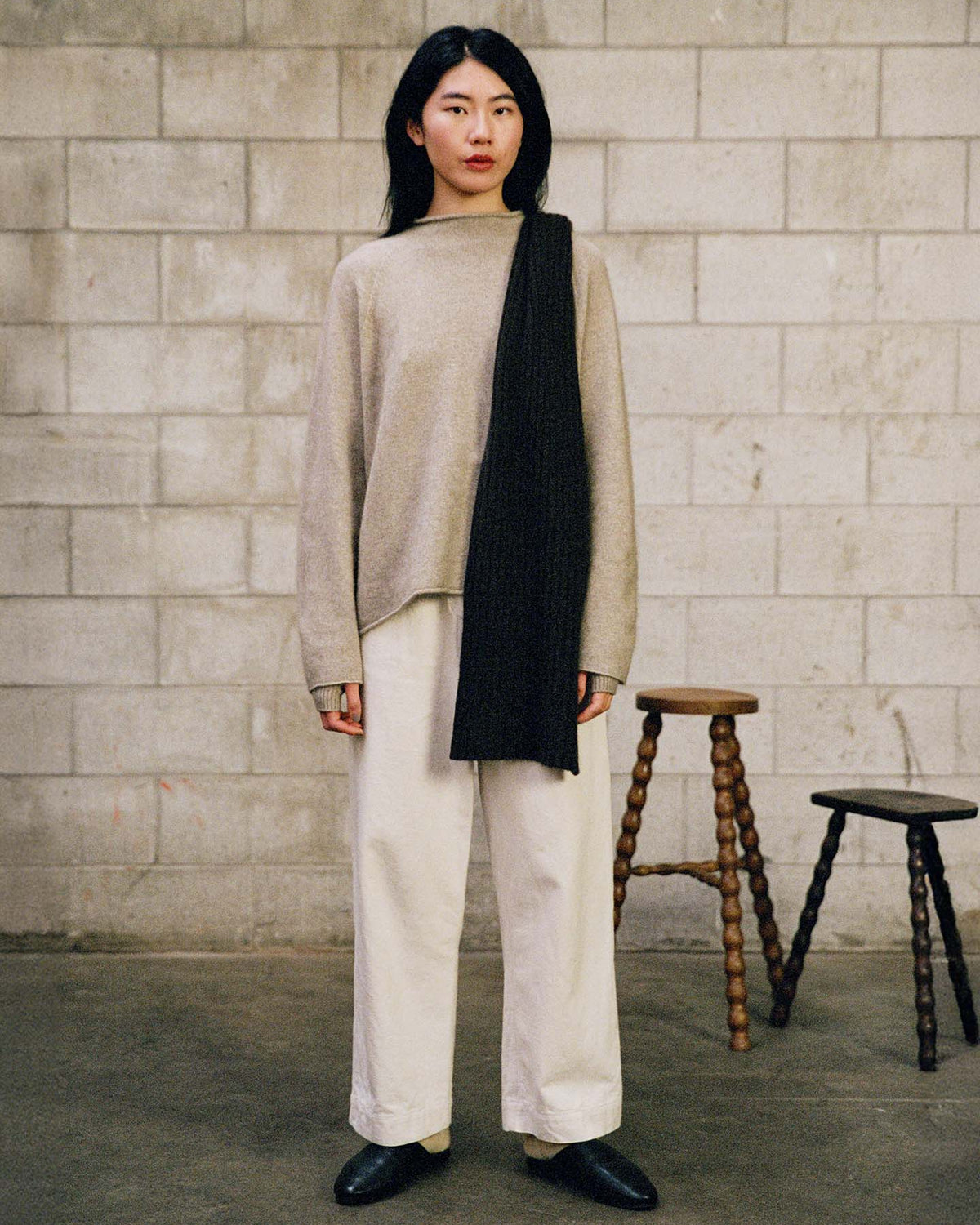 A person stands indoors against a concrete block wall, wearing the Eclipse Knit – Natural sweater, white pants, black shoes, and a black scarf. Two wooden stools are positioned behind them.