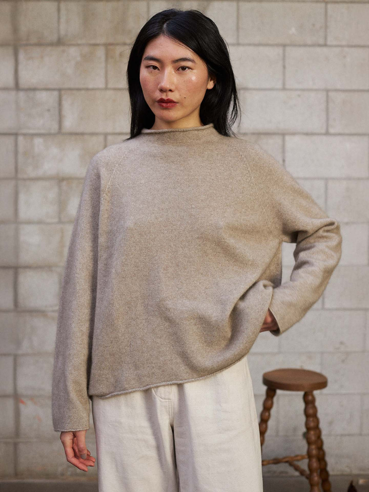 A person with long black hair wears the Eclipse Knit – Natural in a relaxed fit and light pants, standing indoors against a concrete block wall with a wooden stool in the background.