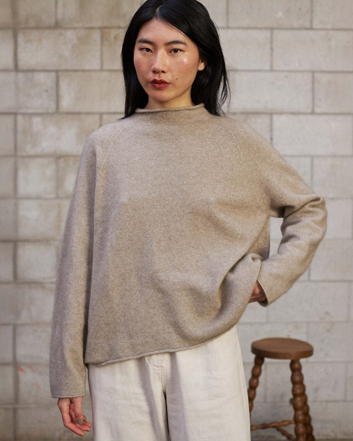 A person with long black hair wears the Eclipse Knit – Natural in a relaxed fit and light pants, standing indoors against a concrete block wall with a wooden stool in the background.