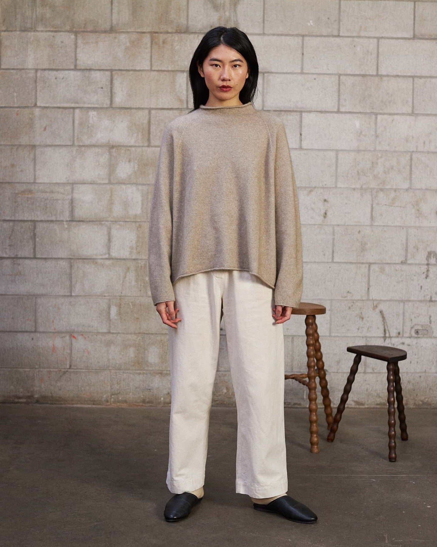 A person stands indoors against a concrete block wall, wearing the Eclipse Knit – Natural sweater with white pants and black shoes. Two wooden stools are placed behind them.