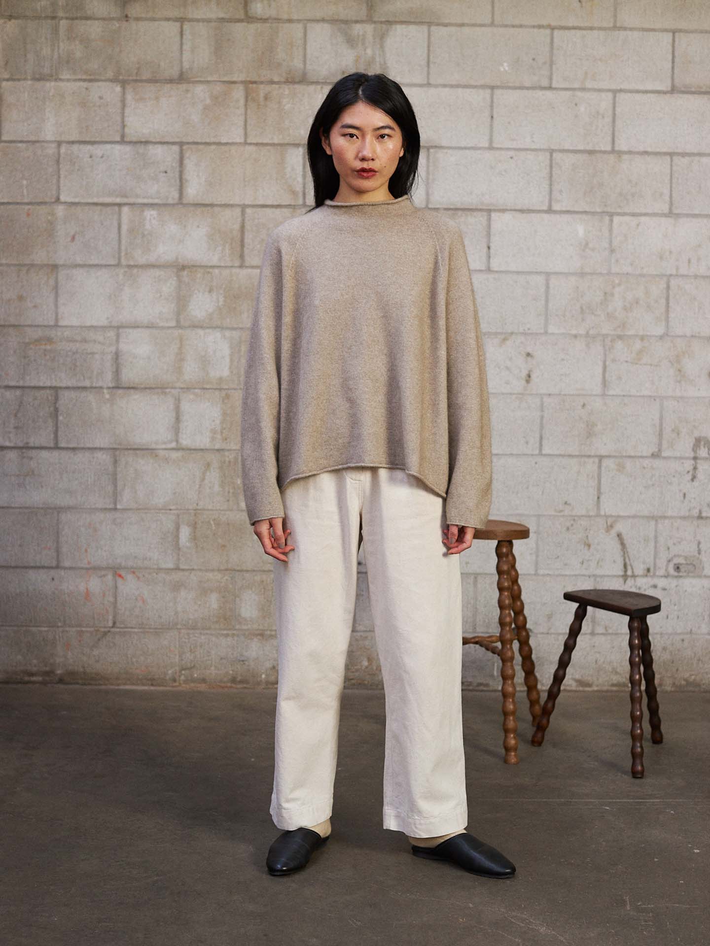 A person stands indoors against a concrete block wall, wearing the Eclipse Knit – Natural sweater with white pants and black shoes. Two wooden stools are placed behind them.