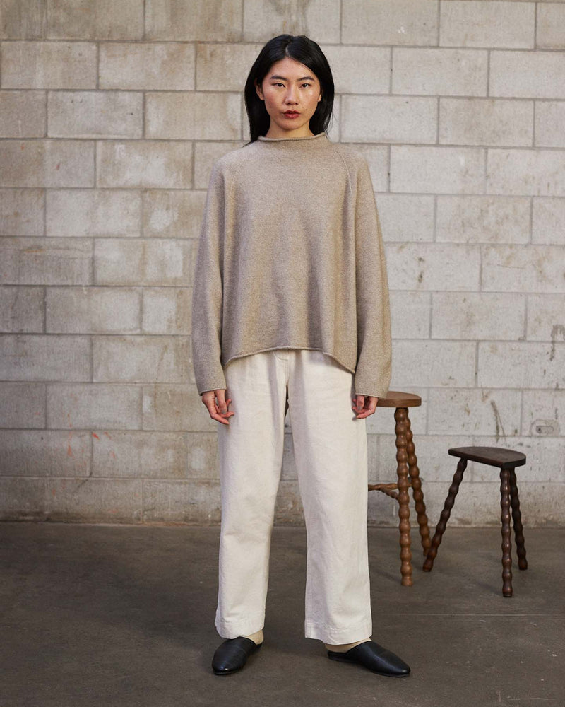 A person stands indoors against a concrete block wall, wearing the Eclipse Knit – Natural sweater with white pants and black shoes. Two wooden stools are placed behind them.