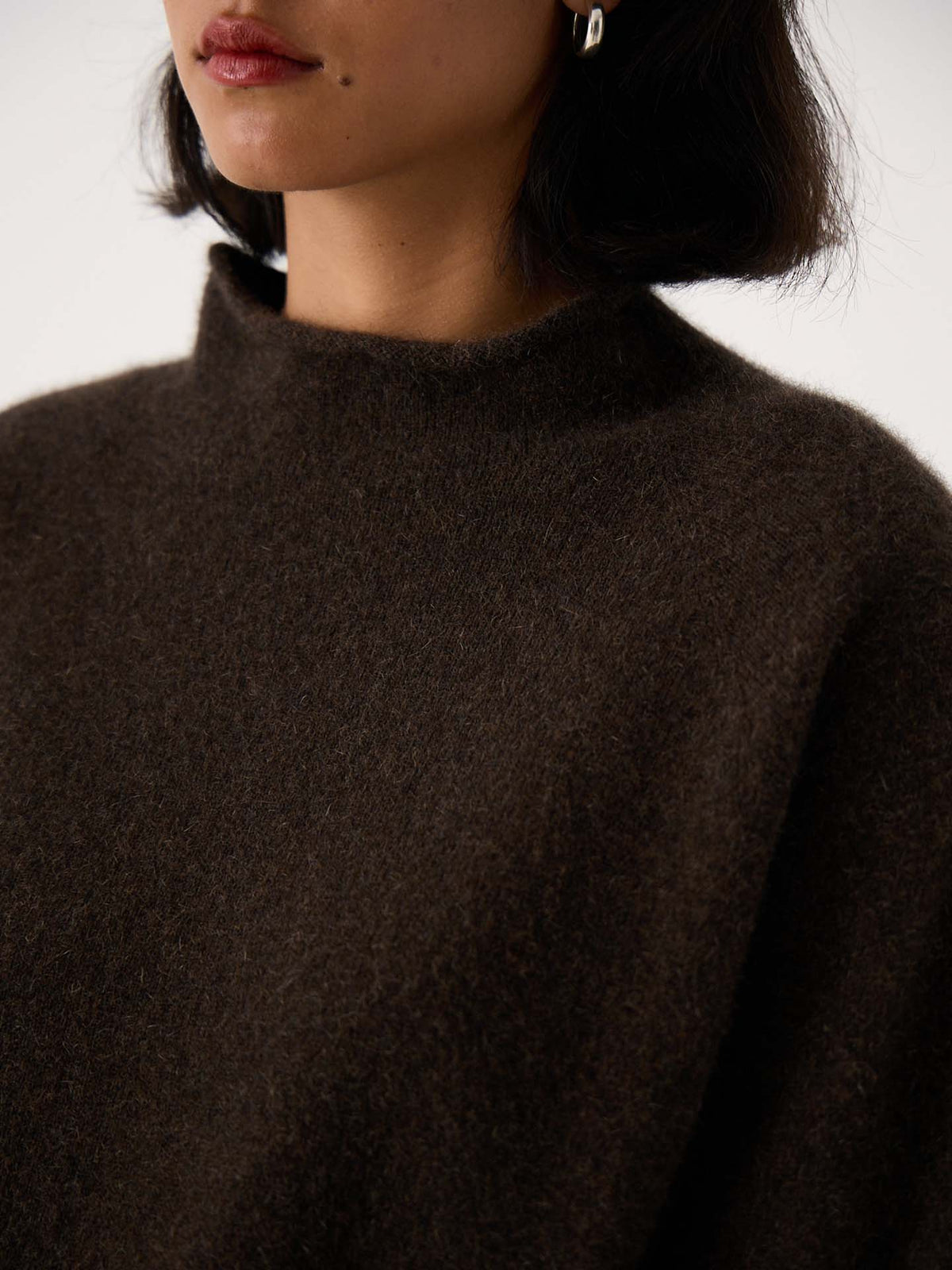 A petite woman with short dark hair wears the Eclipse Knit – Truffle, a relaxed, oversized high-neck fuzzy sweater in dark brown. She also has silver hoop earrings. Only her lower face and upper torso are shown.