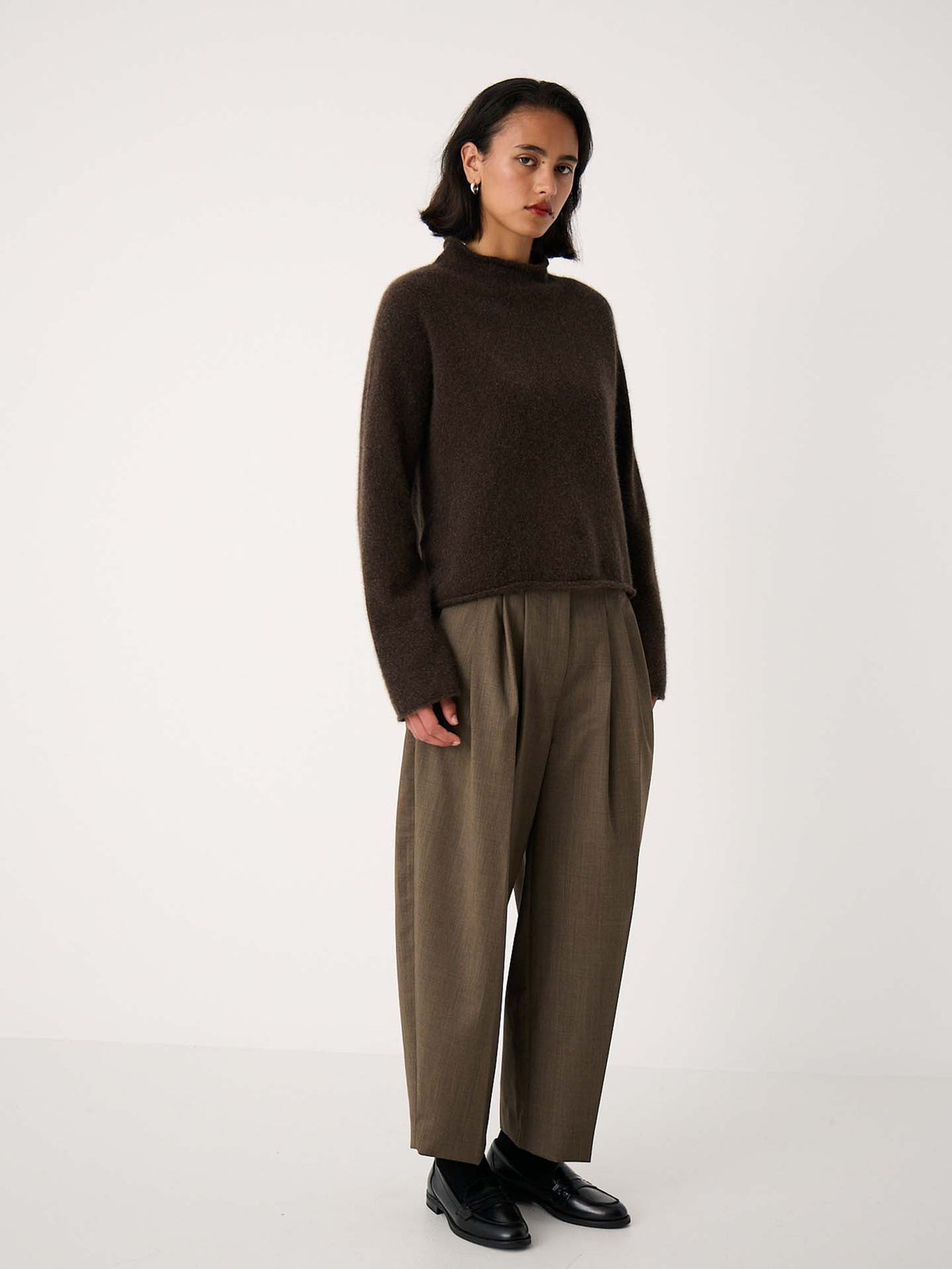 A petite woman stands against a plain white background, wearing the Eclipse Knit – Truffle sweater paired with loose brown trousers and oversized black shoes.