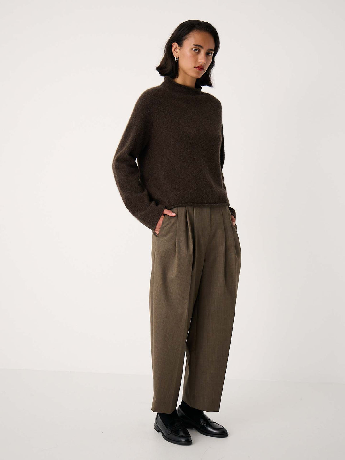 A petite woman stands against a plain background, wearing the Eclipse Knit – Truffle sweater in an oversized fit, paired with loose olive trousers and black shoes, her hands tucked in her pockets for a relaxed look.
