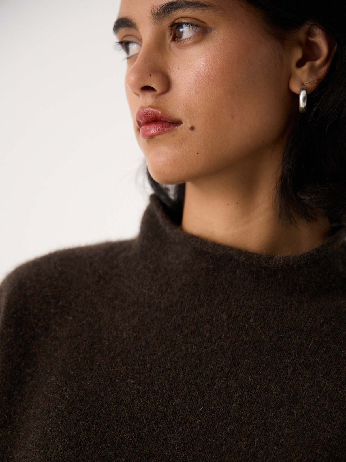 A petite woman with short dark hair wears the Eclipse Knit – Truffle, a relaxed, oversized dark brown sweater, and silver hoop earrings while looking to the side against a plain background.
