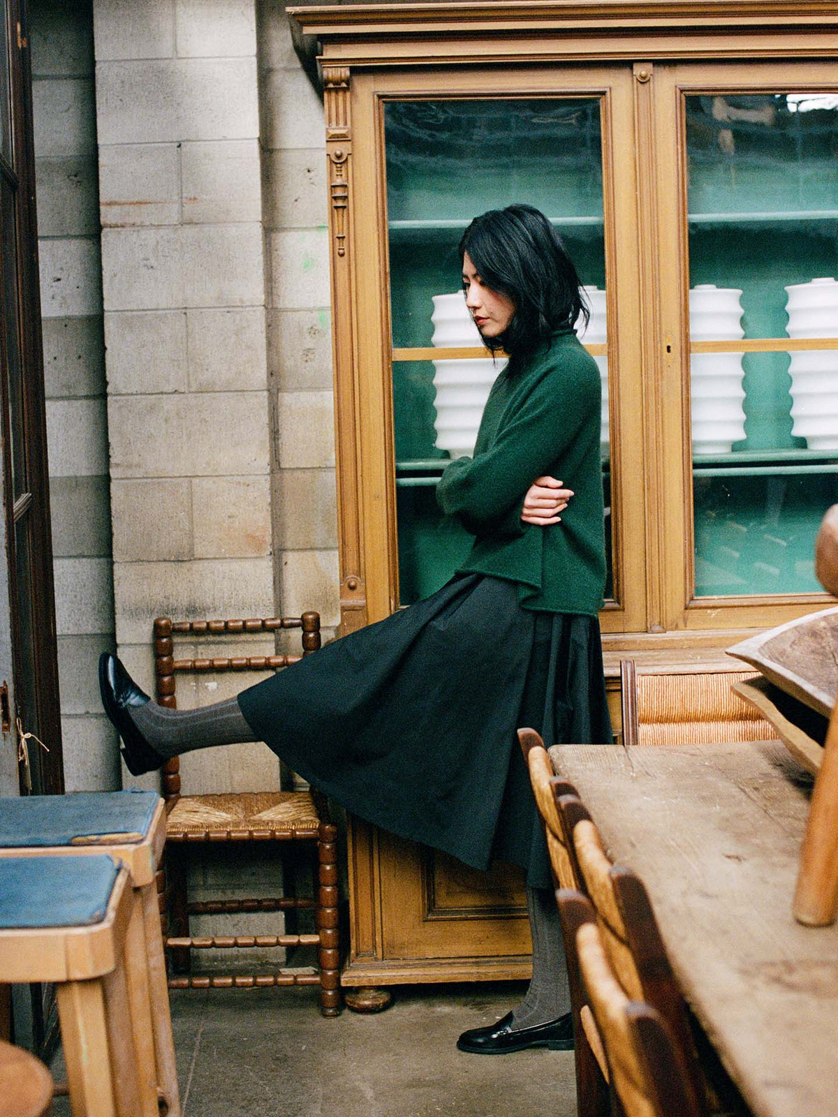 A petite, dark-haired person stands indoors, arms crossed and one leg extended, wearing the Eclipse Knit – Verdant Green in a relaxed fit with a black skirt, surrounded by wooden furniture and cabinets.