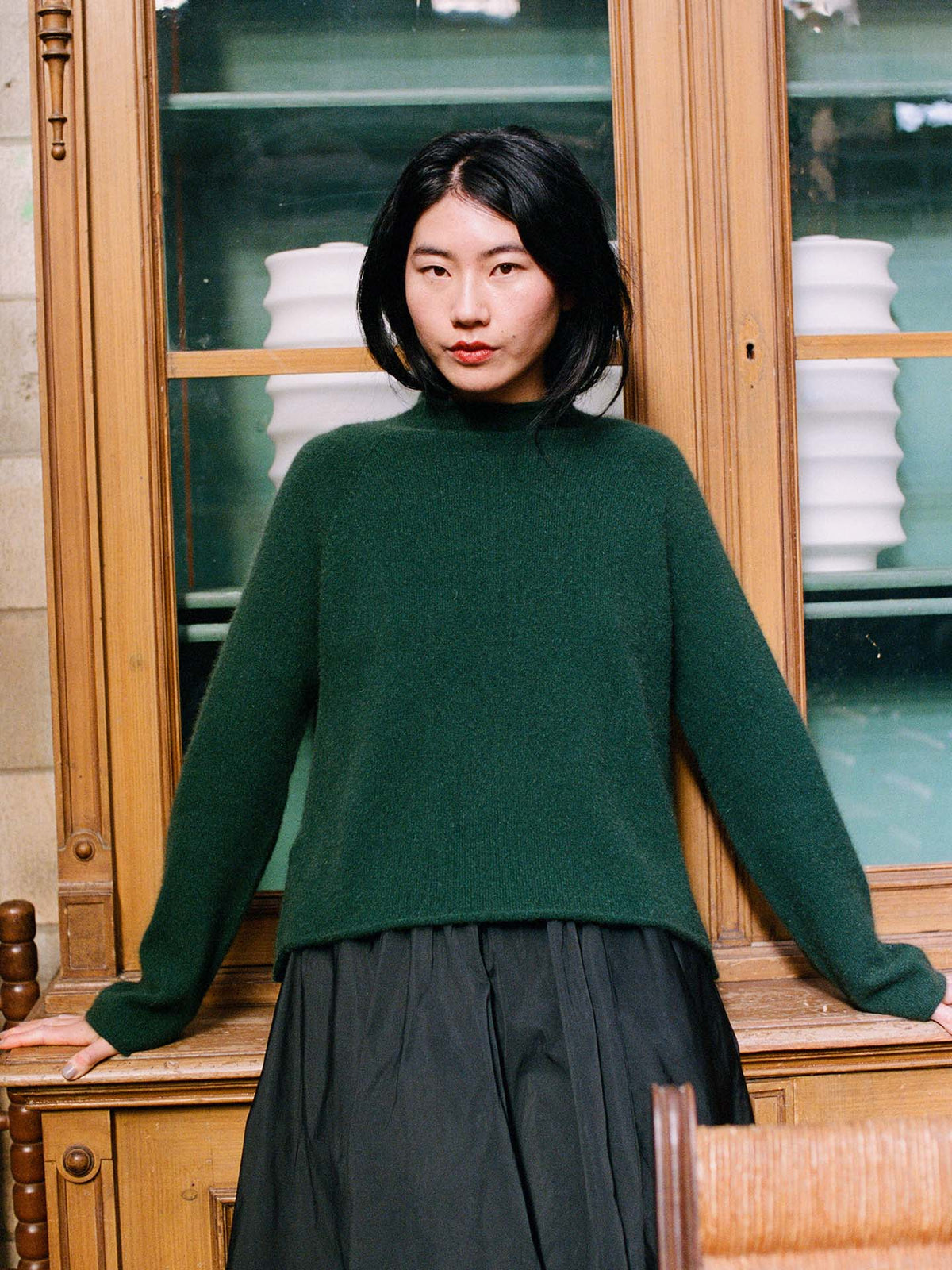 A woman with shoulder-length black hair wears the Eclipse Knit in Verdant Green, a relaxed fit sweater, paired with a black skirt, standing before a wooden cabinet filled with white dishes.