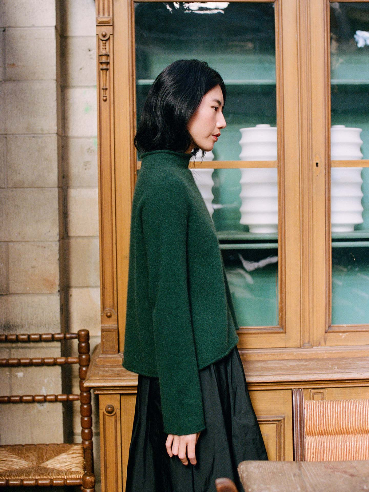 A petite woman with shoulder-length dark hair, wearing the Eclipse Knit – Verdant Green sweater and a black skirt, stands in profile beside a wooden cabinet filled with stacked white dishes.