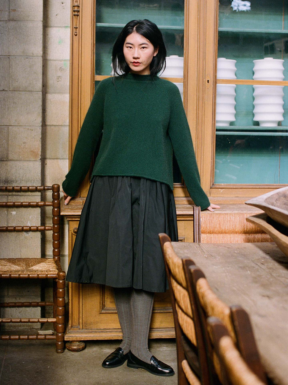 A woman stands indoors by a wooden cabinet, wearing the Eclipse Knit in Verdant Green, paired with a black skirt, gray tights, and black loafers. She faces the camera with her arms at her sides.