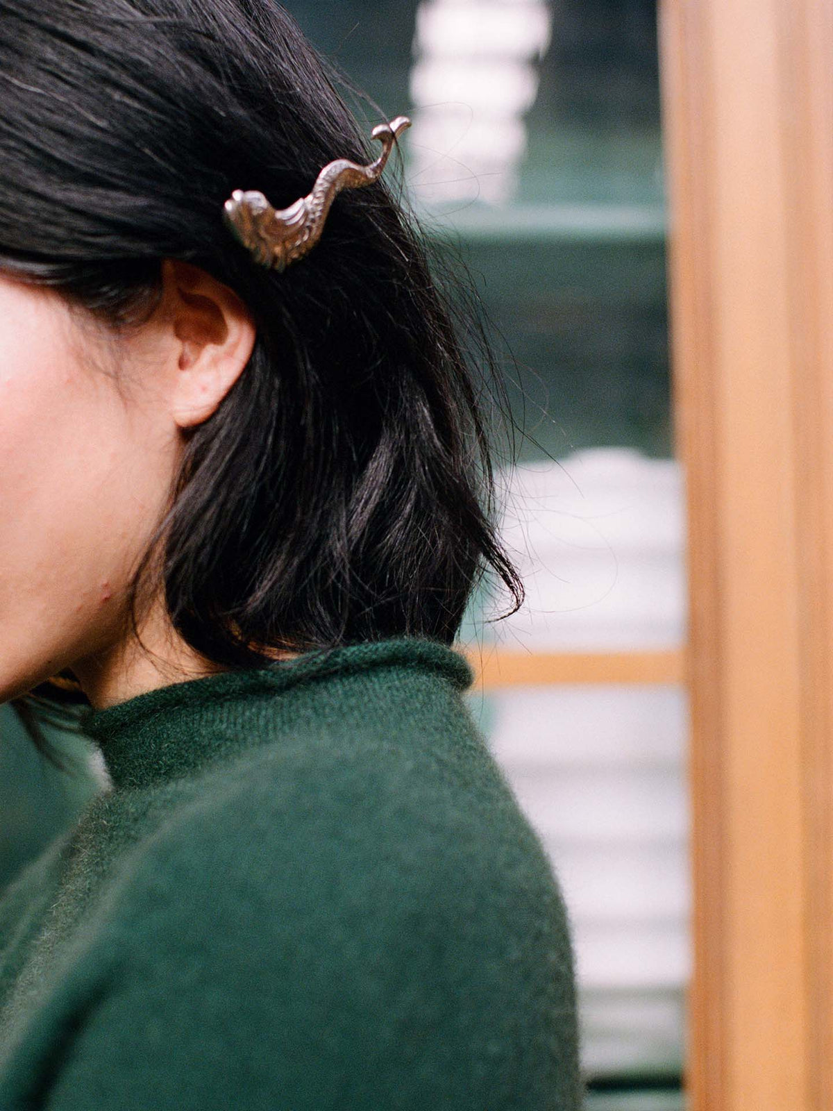 A person with chin-length black hair wears the Eclipse Knit – Verdant Green in a relaxed fit and has a silver bird-shaped hair clip on the left side of their head.