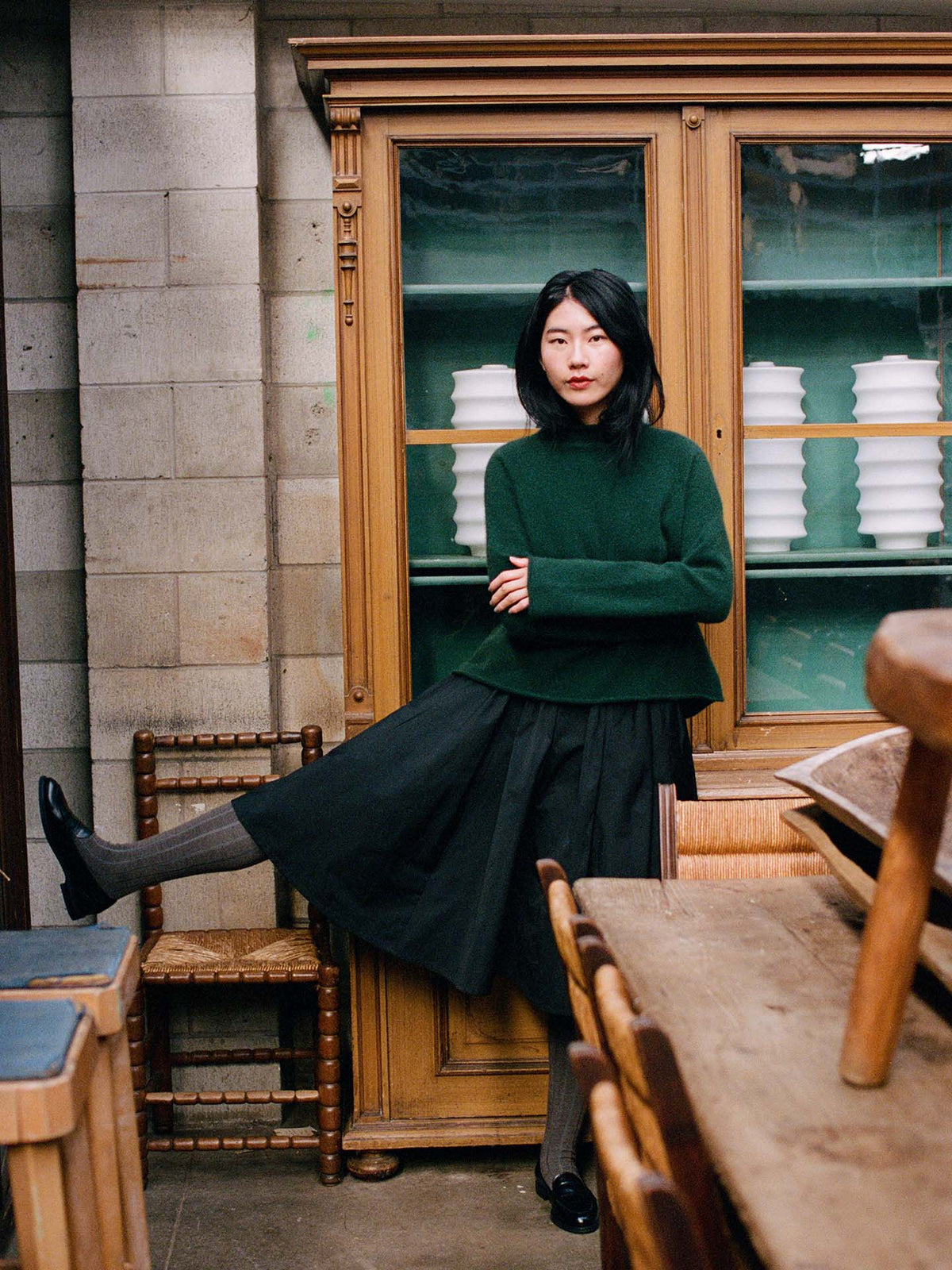 A woman in the Eclipse Knit – Verdant Green sweater and a black skirt stands indoors, one leg raised on a chair, in front of a wooden cabinet with stacked white dishes.