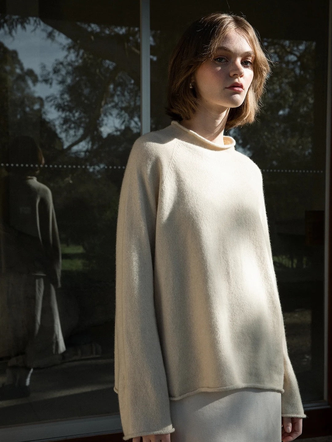 A young person with short hair, radiating a laid-back vibe, is seen wearing the oversized "Eclipse Knit – Creme" by Francie. They stand in front of a reflective glass surface, showcasing trees in the background.