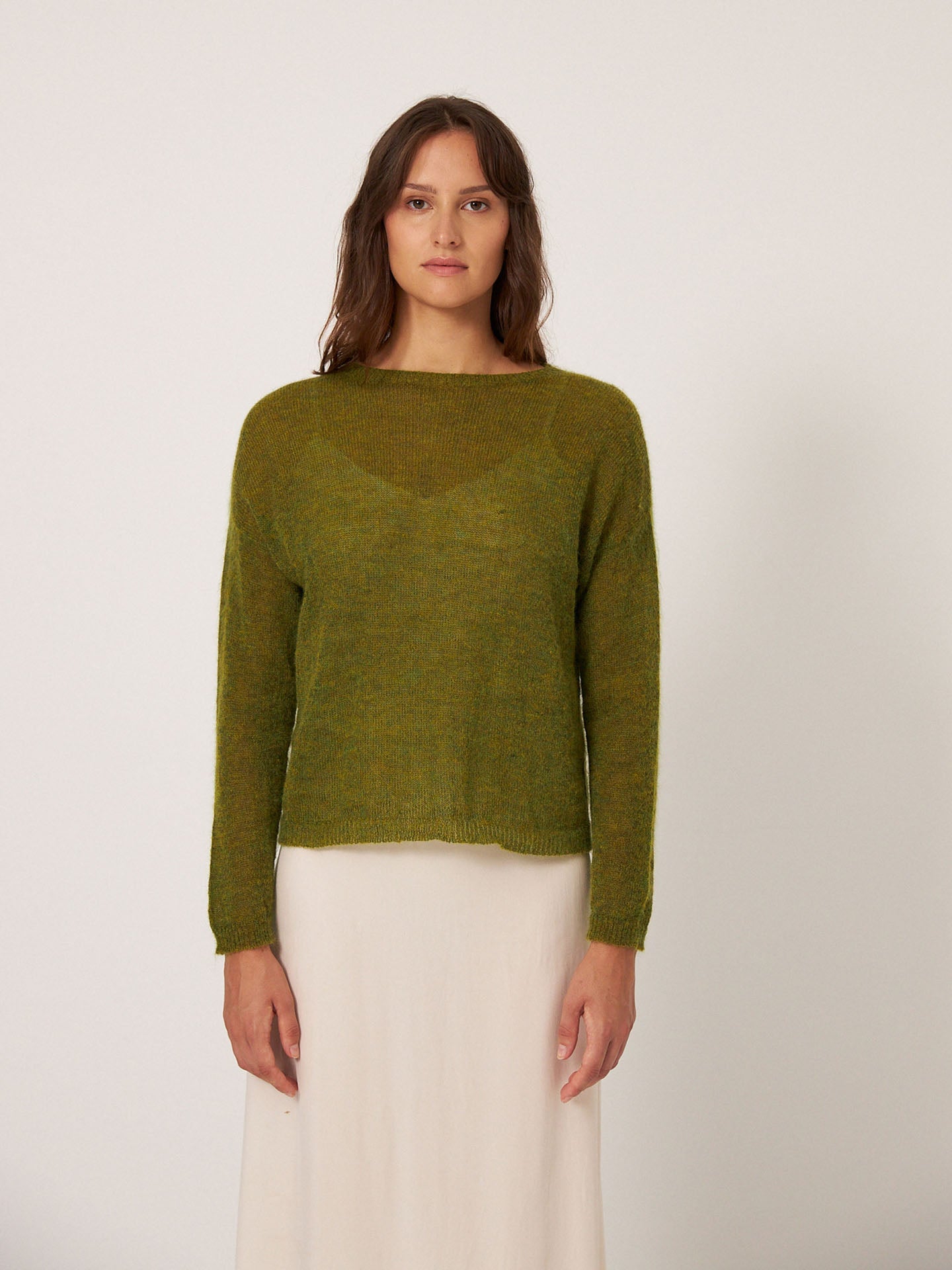 A person with long brown hair, styled in a relaxed and oversized "Feather Knit – Peridot Green" sweater by Francie, stands against a plain background, paired with a white skirt.