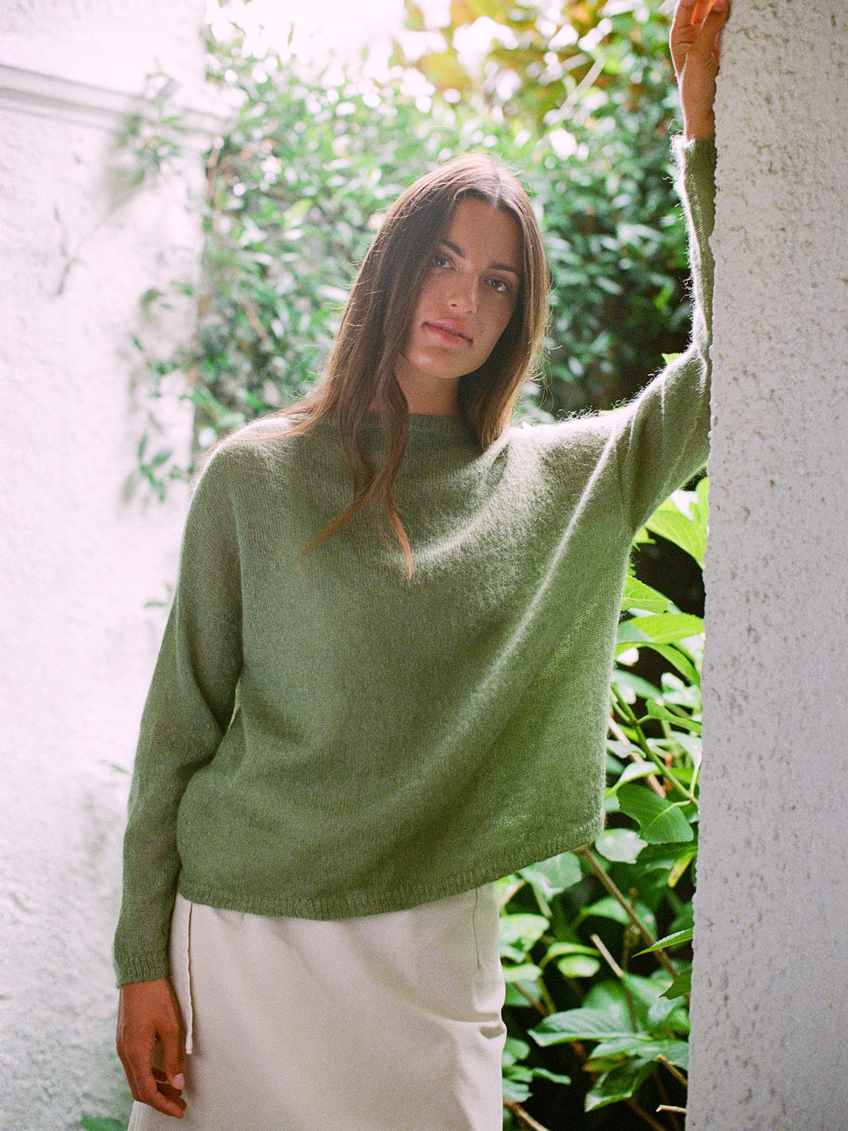 A woman wears the Feather Knit – Clover, an oversized green sweater, paired with a cream skirt as she stands against a white wall with greenery in the background.