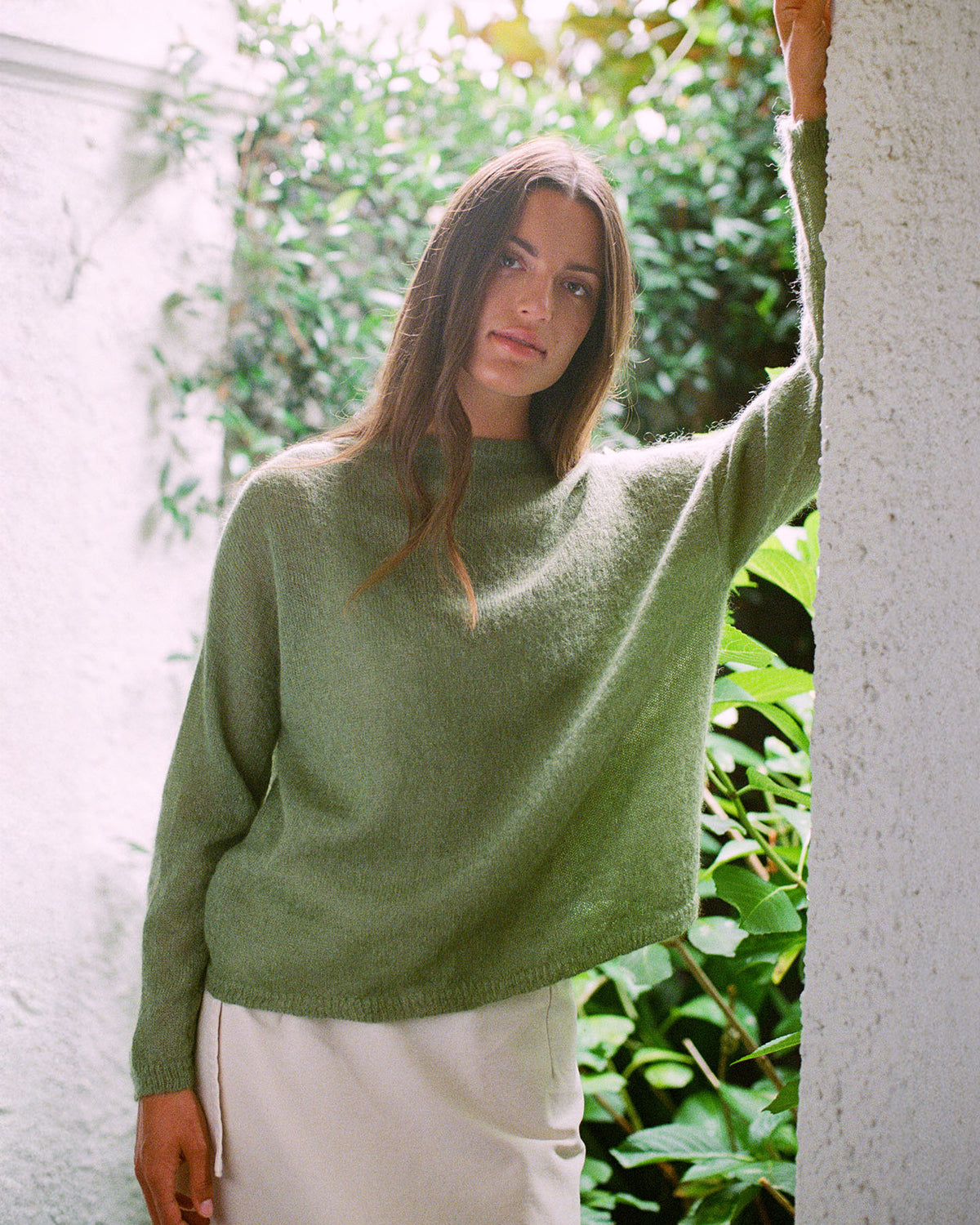 A woman wears the Feather Knit – Clover, an oversized green sweater, paired with a cream skirt as she stands against a white wall with greenery in the background.
