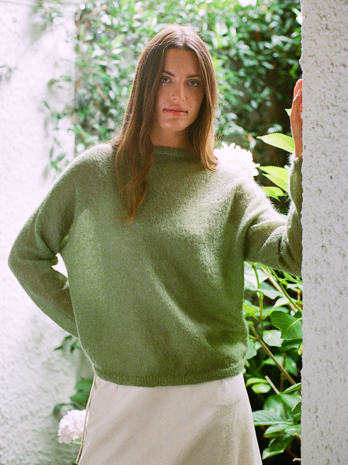 A woman with long brown hair wears the Feather Knit – Clover sweater and a light skirt, standing before a lush, green leafy backdrop.