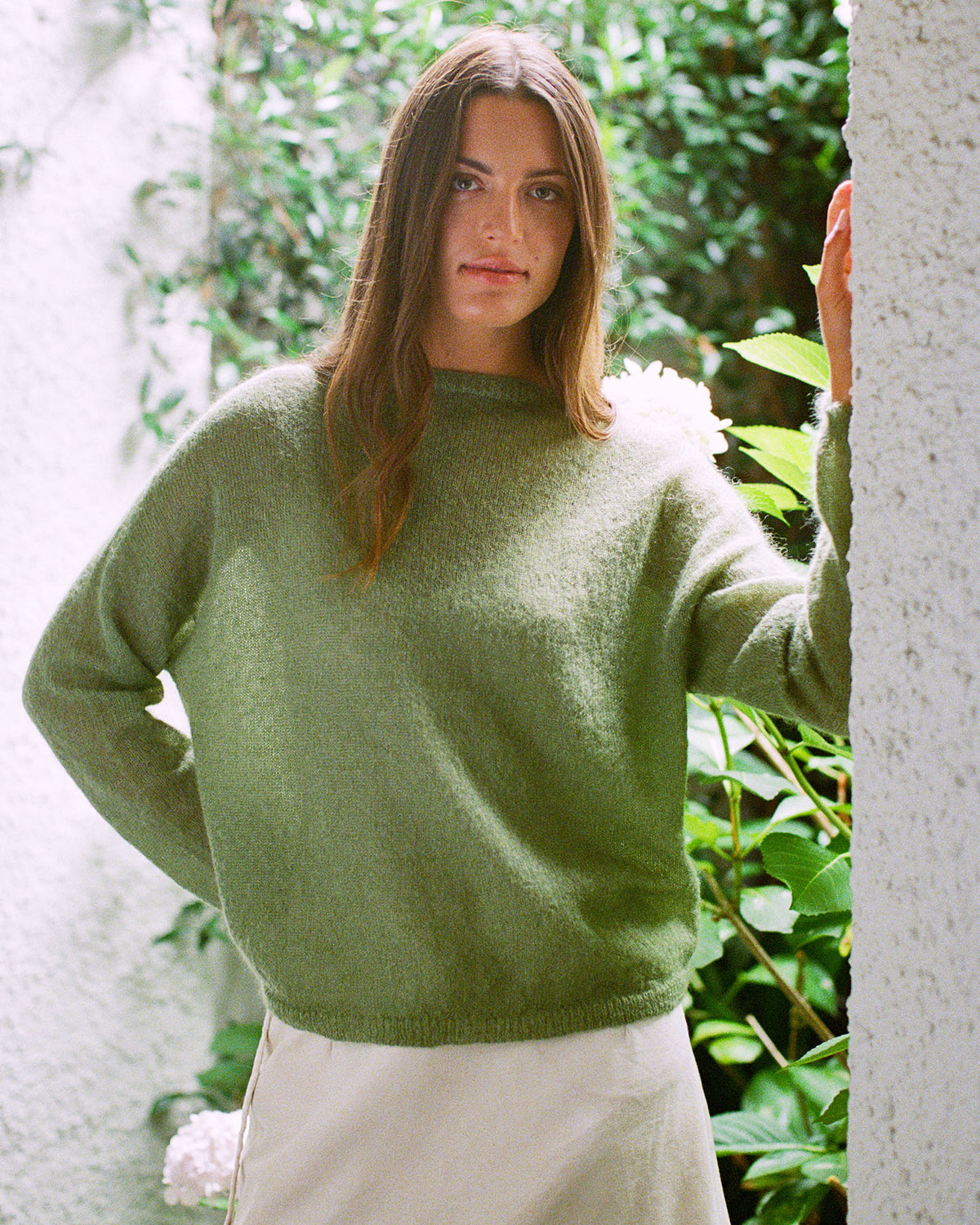 A woman with long brown hair wears the Feather Knit – Clover sweater and a light skirt, standing before a lush, green leafy backdrop.