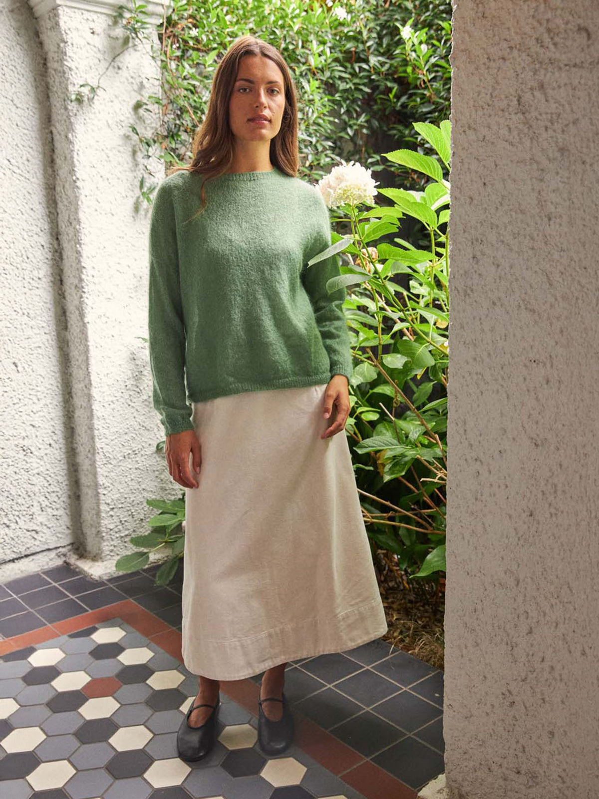 A woman stands on a tiled floor near greenery, wearing the Feather Knit – Clover oversized green sweater, a white ankle-length skirt, and black shoes.