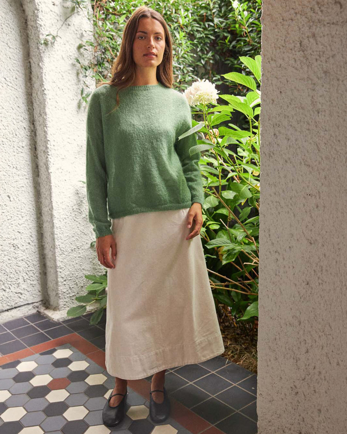A woman stands on a tiled floor near greenery, wearing the Feather Knit – Clover oversized green sweater, a white ankle-length skirt, and black shoes.