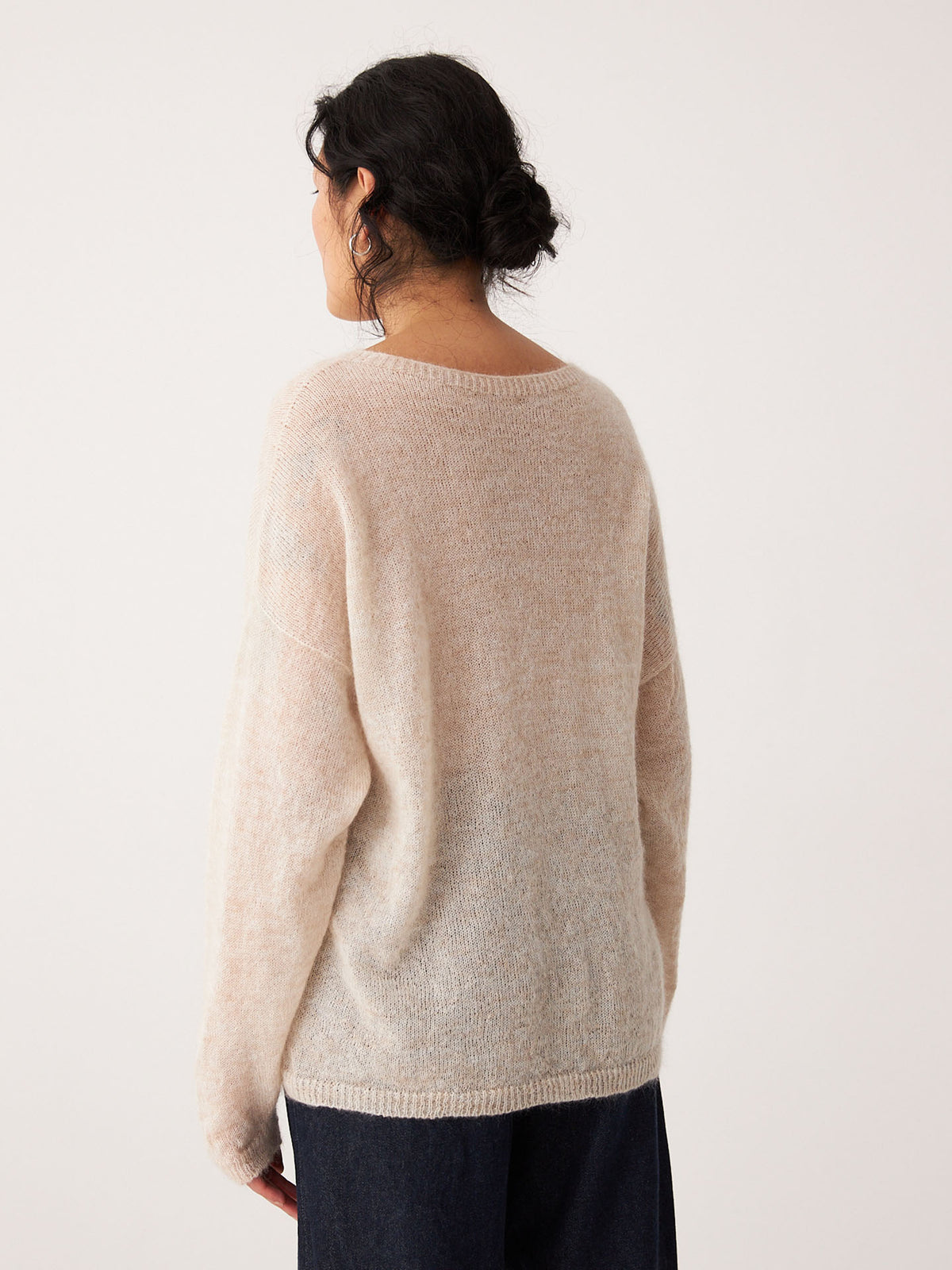 A person with dark hair in a low bun is seen from the back, wearing the Feather Knit – Creme, a relaxed fit, light beige oversized sweater, paired with dark pants against a plain light background.