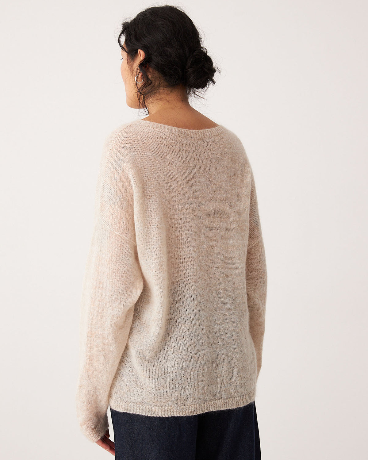 A person with dark hair in a low bun is seen from the back, wearing the Feather Knit – Creme, a relaxed fit, light beige oversized sweater, paired with dark pants against a plain light background.