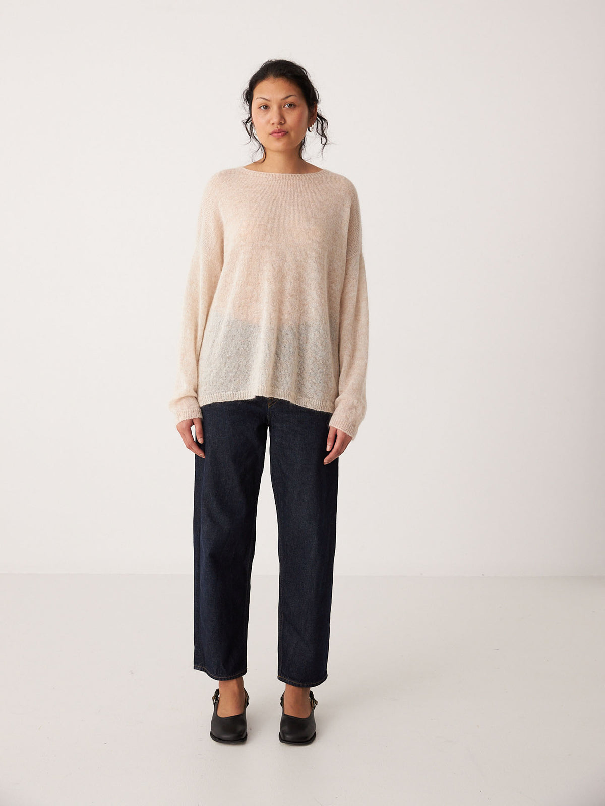 A woman stands facing forward against a plain white background, wearing a relaxed fit Feather Knit – Creme sweater, dark wide-leg jeans, and black shoes.