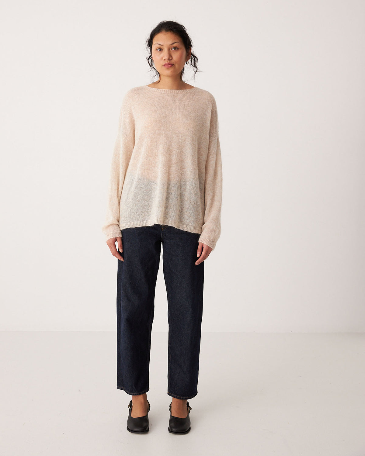 A woman stands facing forward against a plain white background, wearing a relaxed fit Feather Knit – Creme sweater, dark wide-leg jeans, and black shoes.