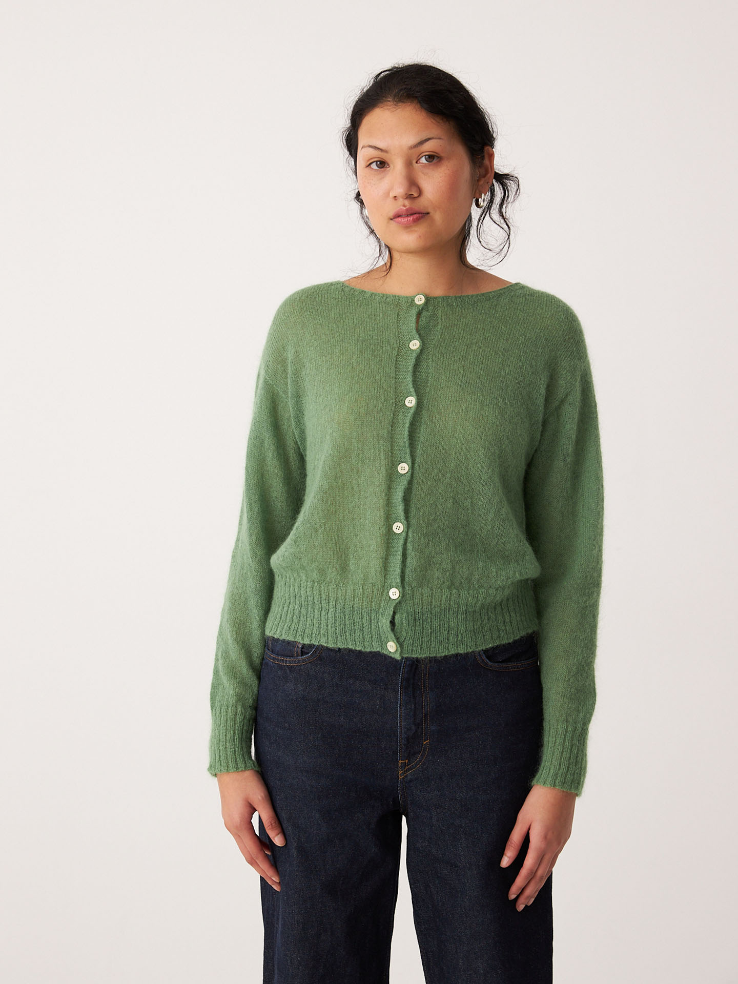 A woman faces the camera wearing the Fleur Cardigan – Clover, a relaxed-fit green button-up, paired with dark blue jeans against a plain white background.