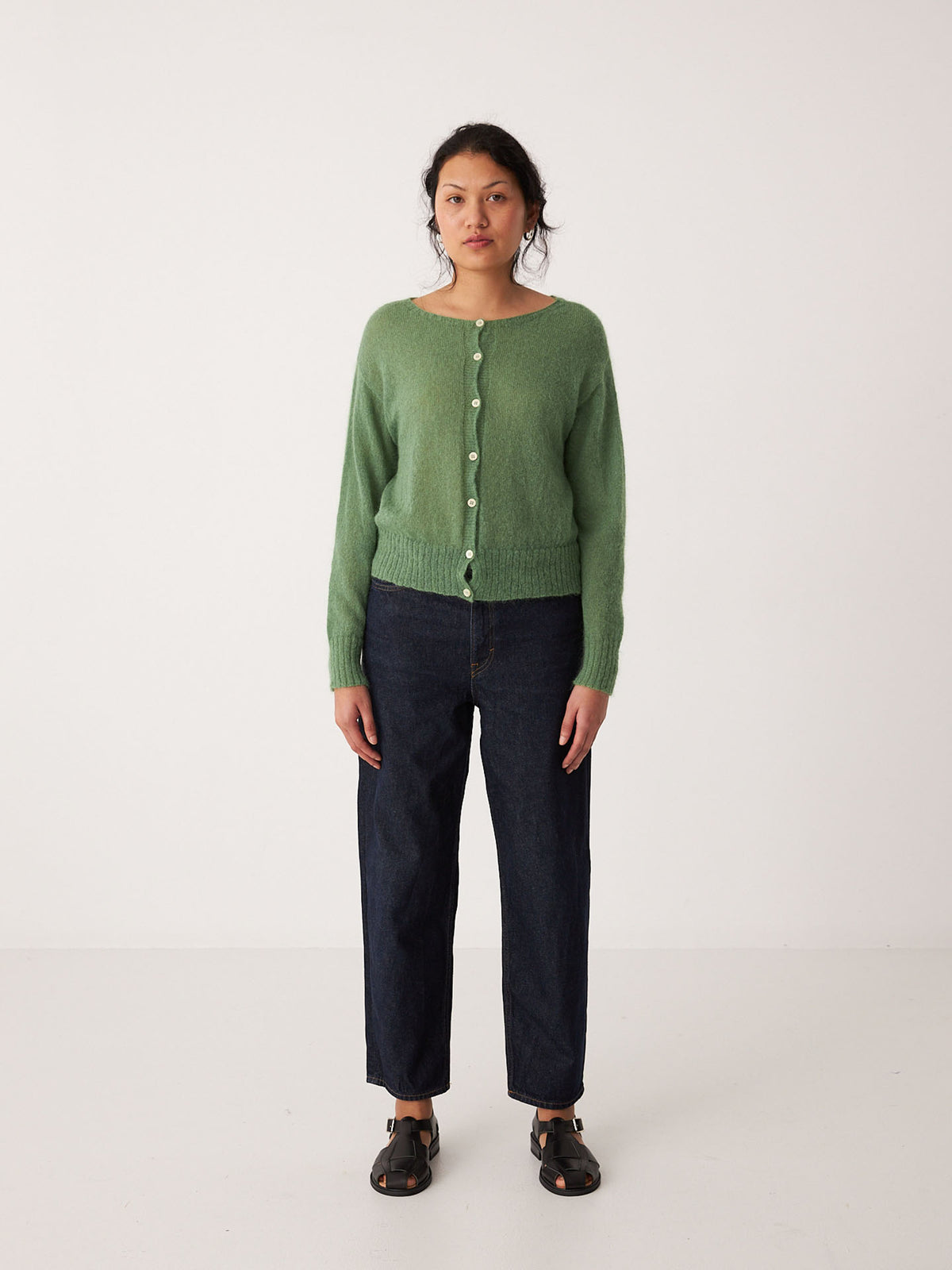 A petite person stands against a plain white background wearing the Fleur Cardigan – Clover, dark blue jeans, and black sandals.