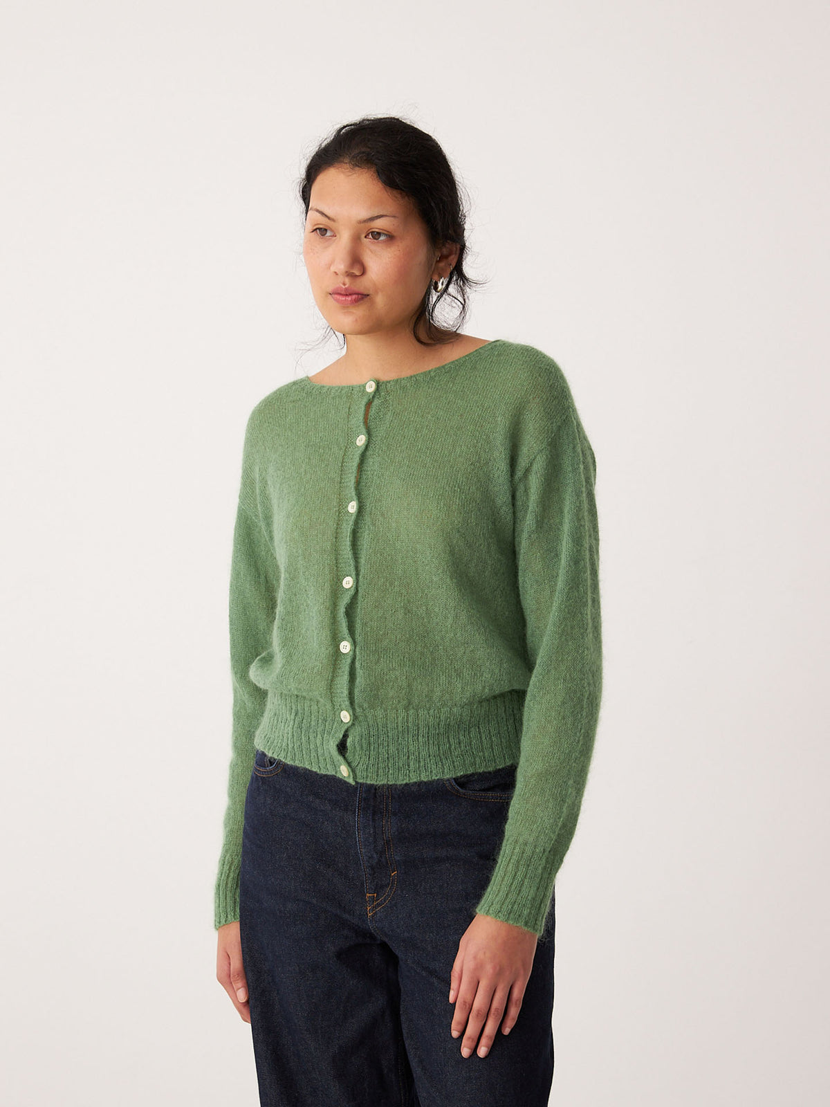 A petite person wears the Fleur Cardigan in Clover, a relaxed fit green button-up, paired with dark blue jeans, standing against a plain white background.