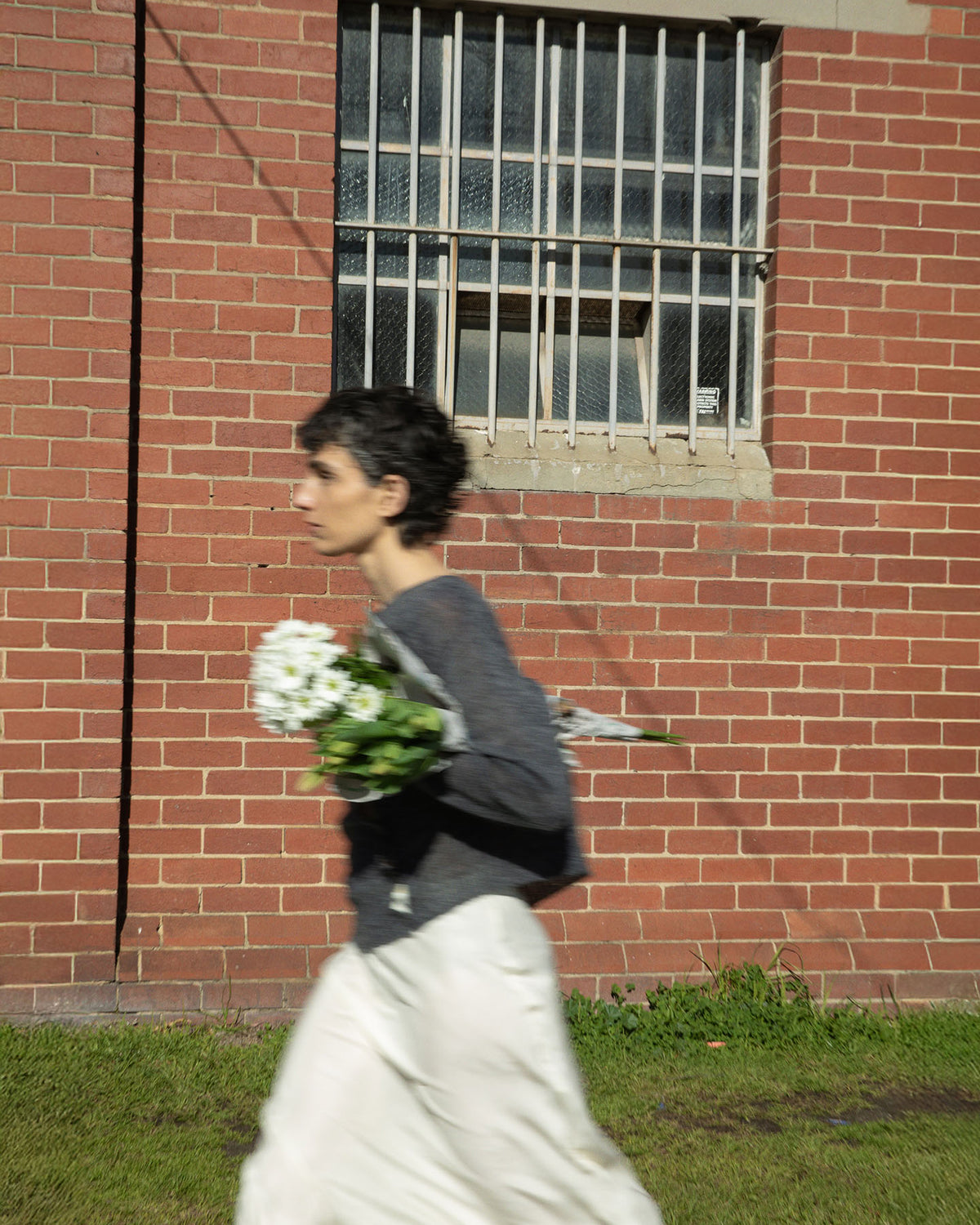 A person in a Fleur Cardigan – Shadow Grey by Francie strolls casually past a red brick building with a barred window, holding flowers.
