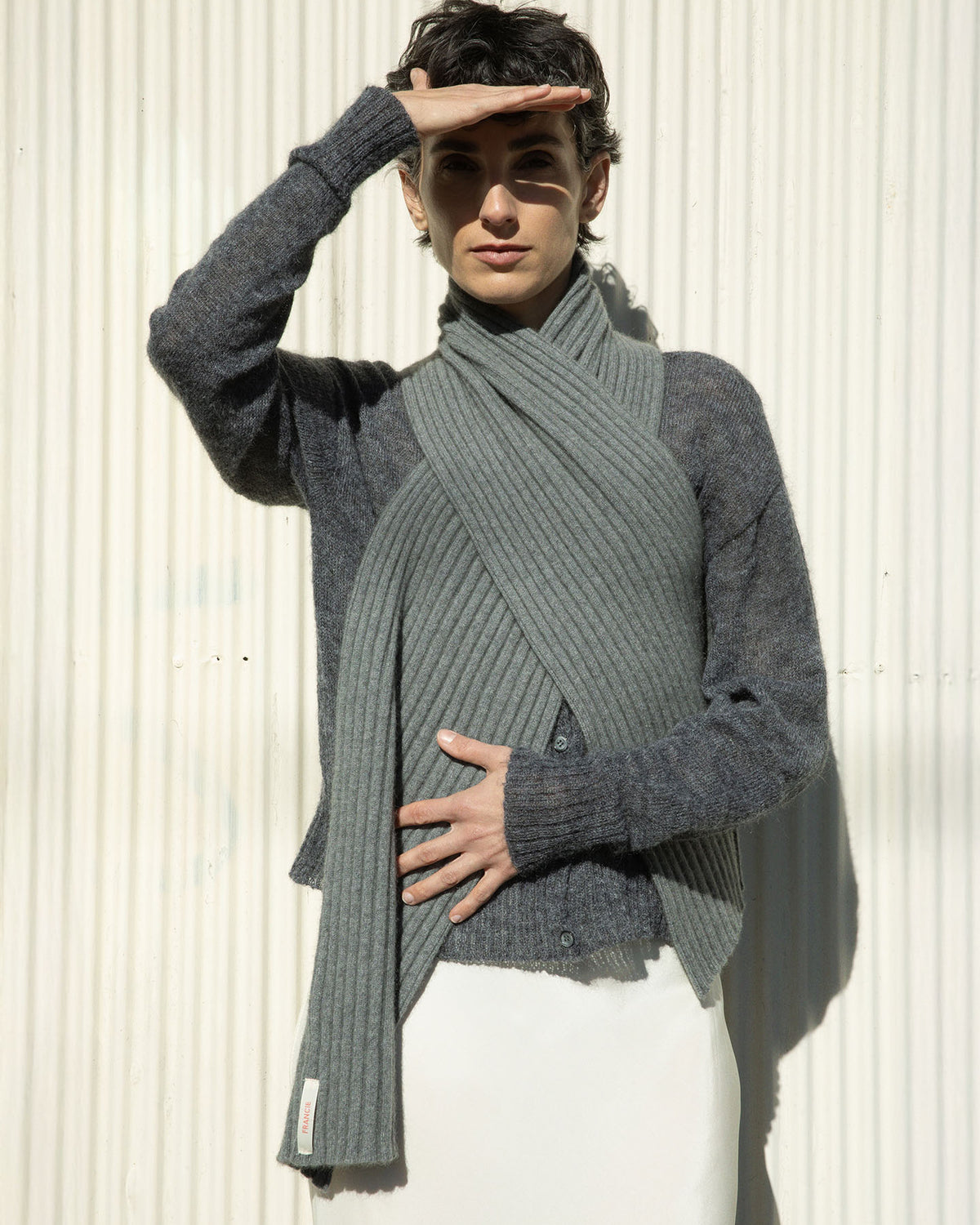A relaxed person in the Francie Fleur Cardigan in Shadow Grey and a scarf leans against a textured wall, shading their eyes with one hand, achieving a chic cropped style.