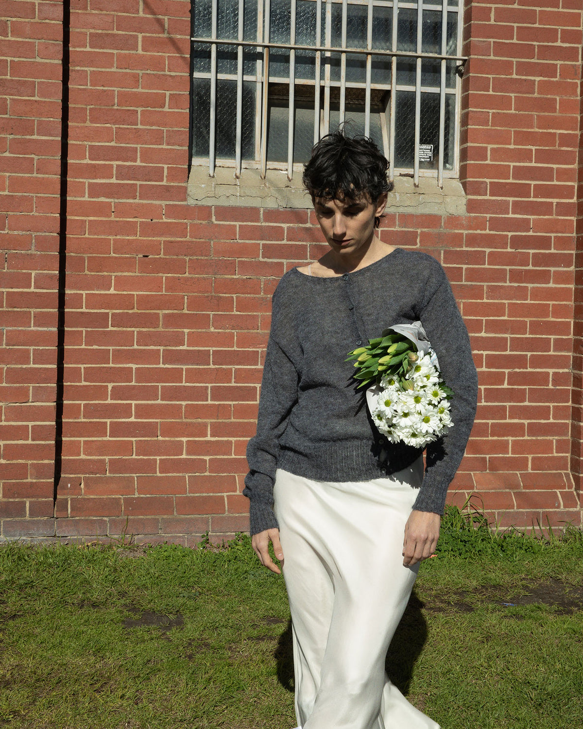 A petite person in a Fleur Cardigan – Shadow Grey by Francie, paired with a white skirt, exudes a relaxed vibe while holding a bouquet of white flowers as she strolls past a brick wall with a barred window.